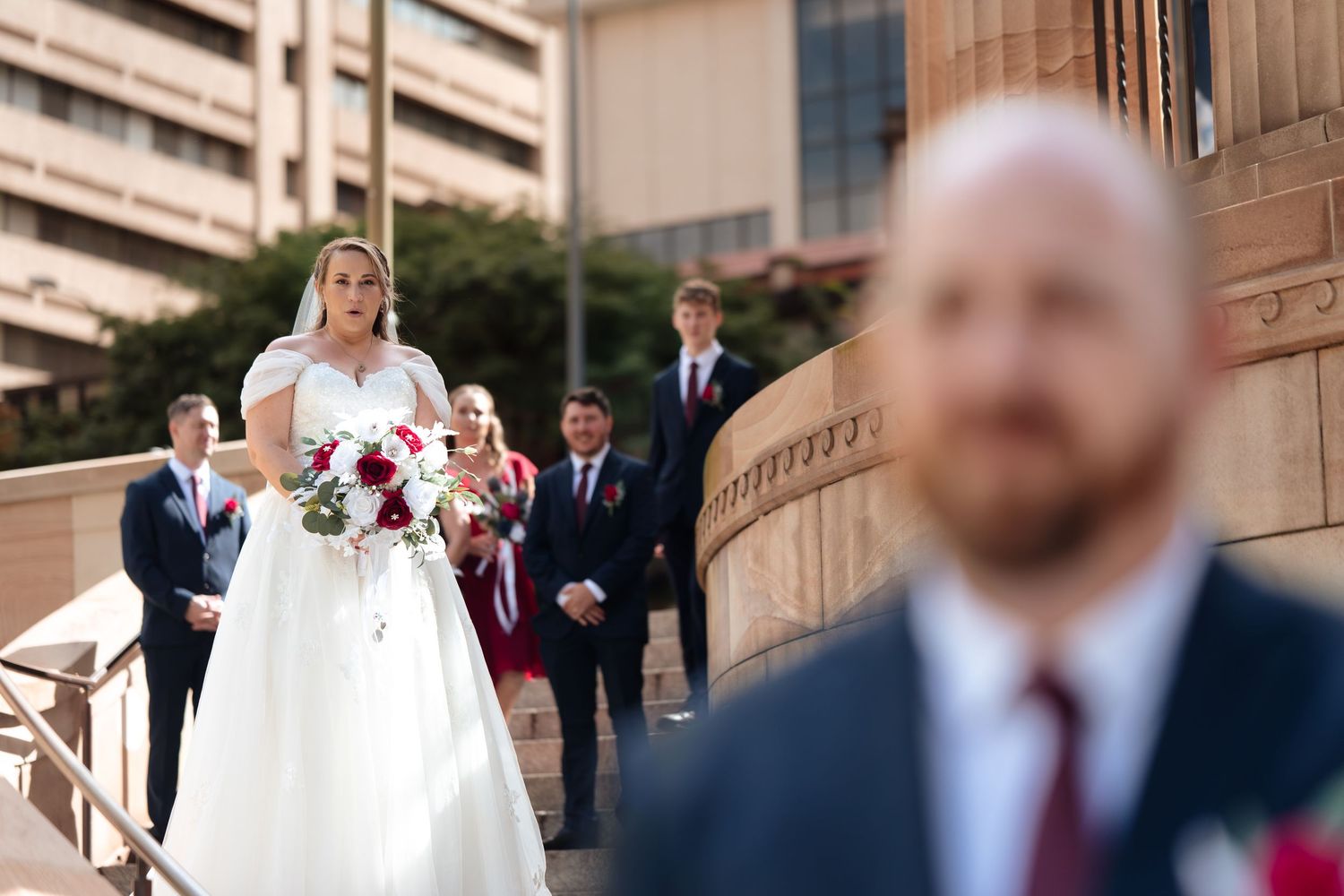 Bride in white gown holding red bouquet walks toward groom during outdoor wedding ceremony with bridal party on steps.