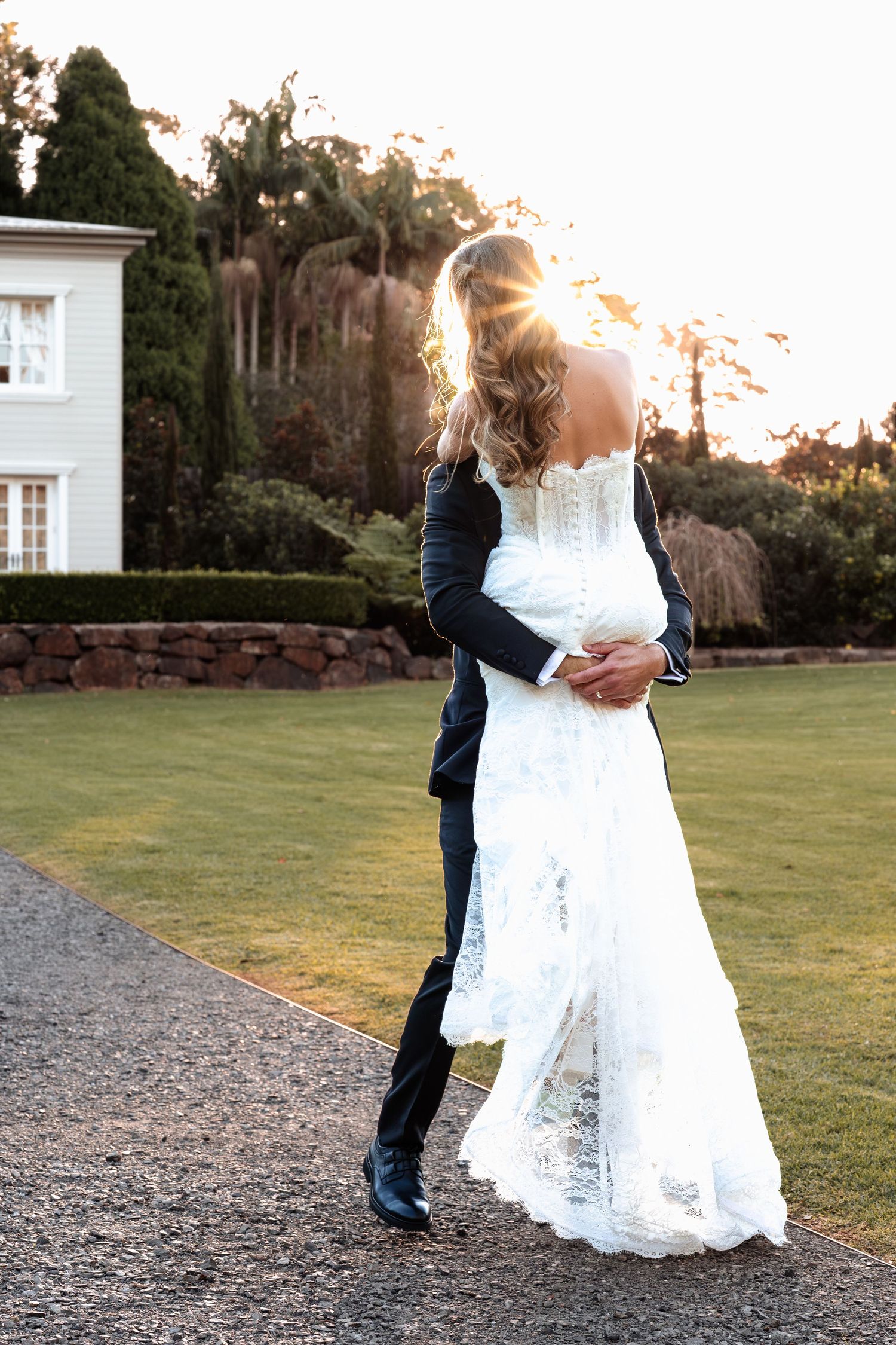 Bride and groom embrace on gravel path at sunset with white wedding dress and formal attire in garden setting.