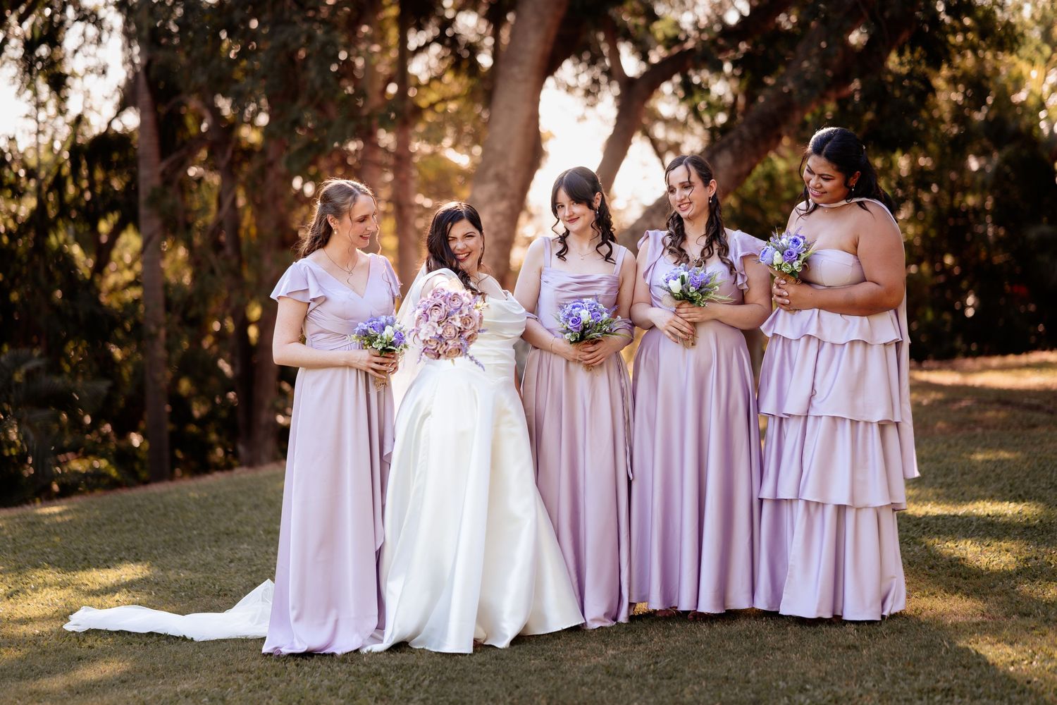 Bride and four bridesmaids in matching blush pink dresses holding bouquets outdoors.