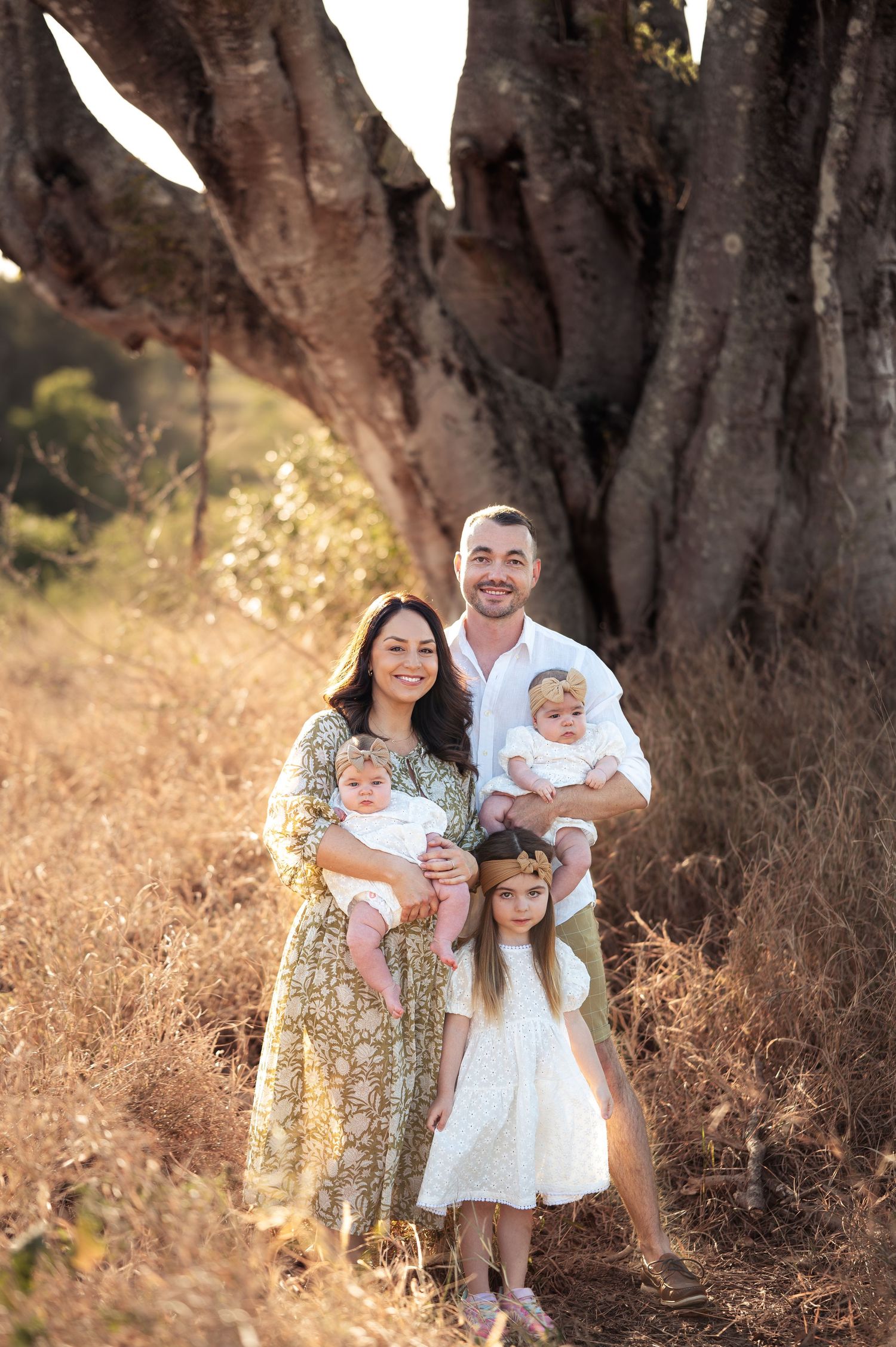 Family wearing white outfits poses together under large tree branches.
