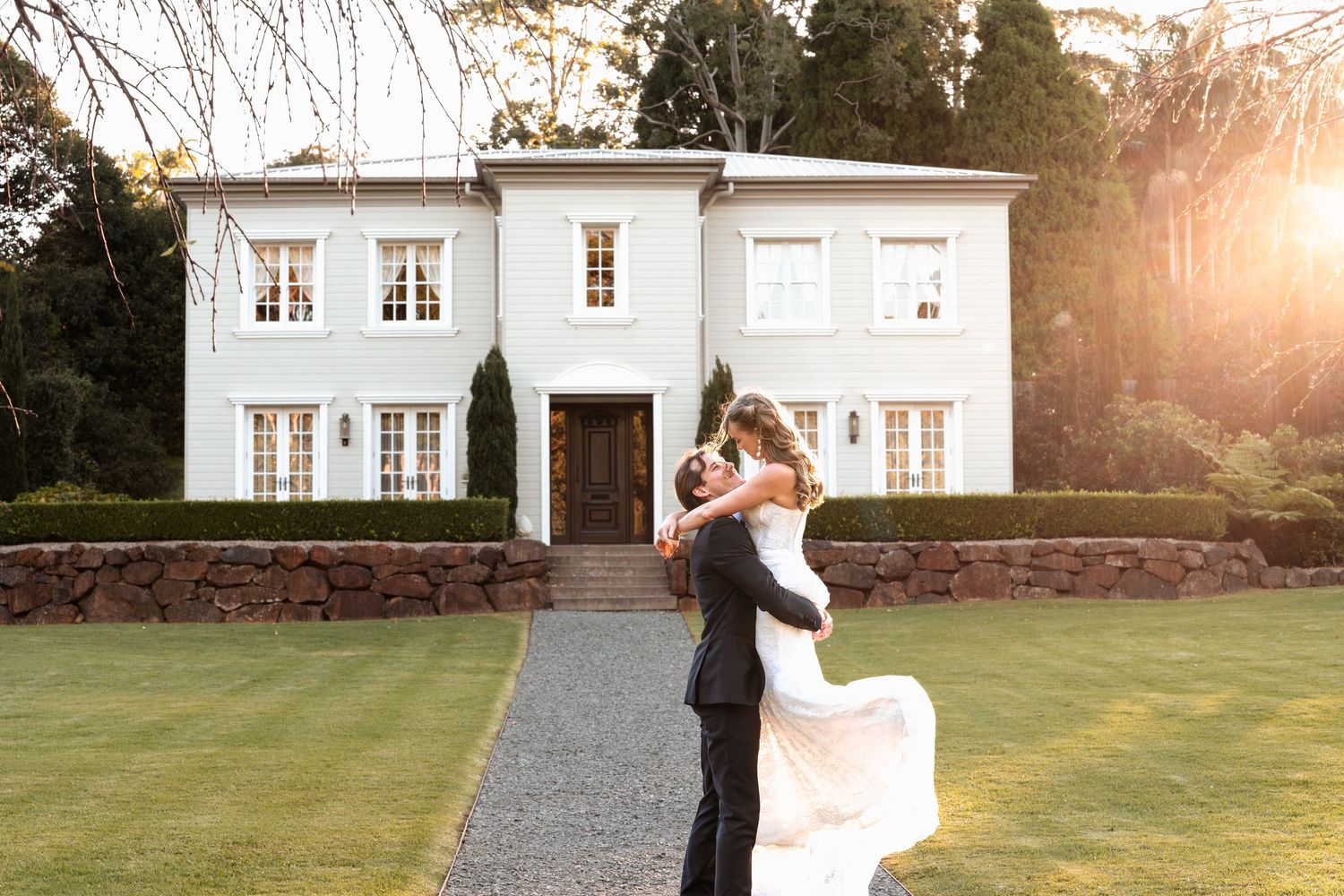 Bride and groom embrace in front of elegant white mansion during golden hour sunset wedding photography.