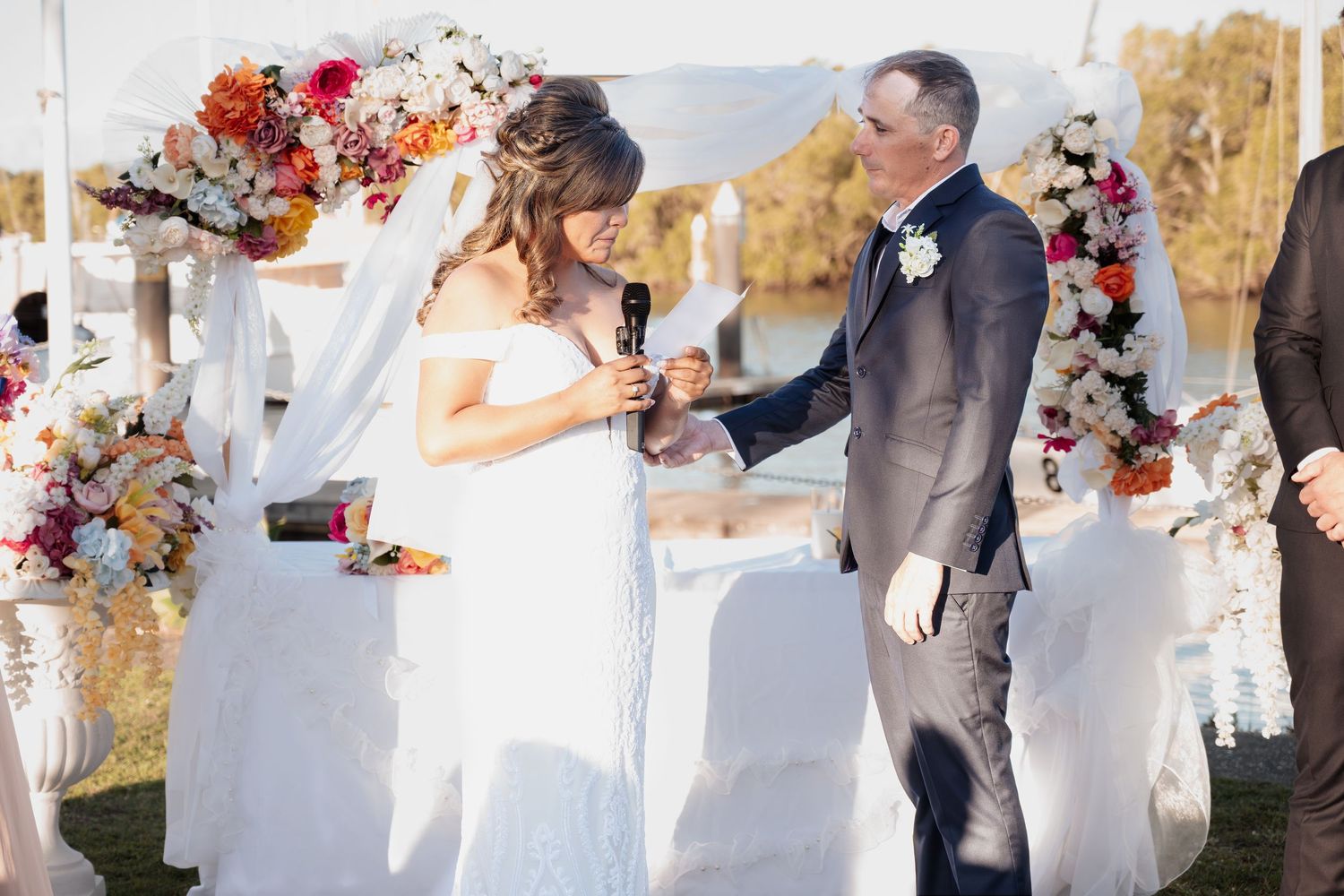 Bride and groom exchanging vows under a floral arch with colorful blooms at an outdoor waterfront wedding ceremony.