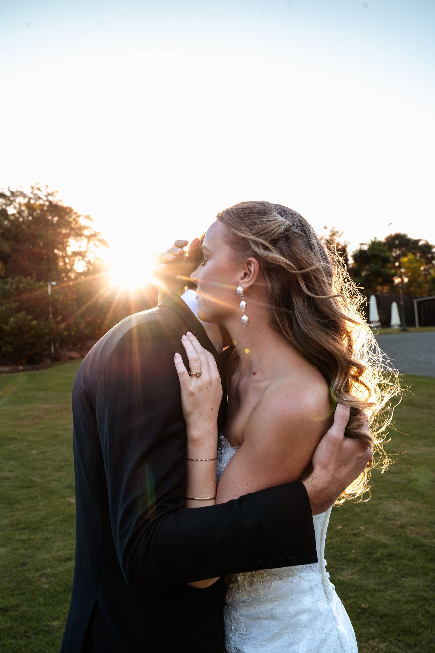 Bride and groom share a romantic sunset kiss outdoors, with golden light glowing behind them on lush green grounds.