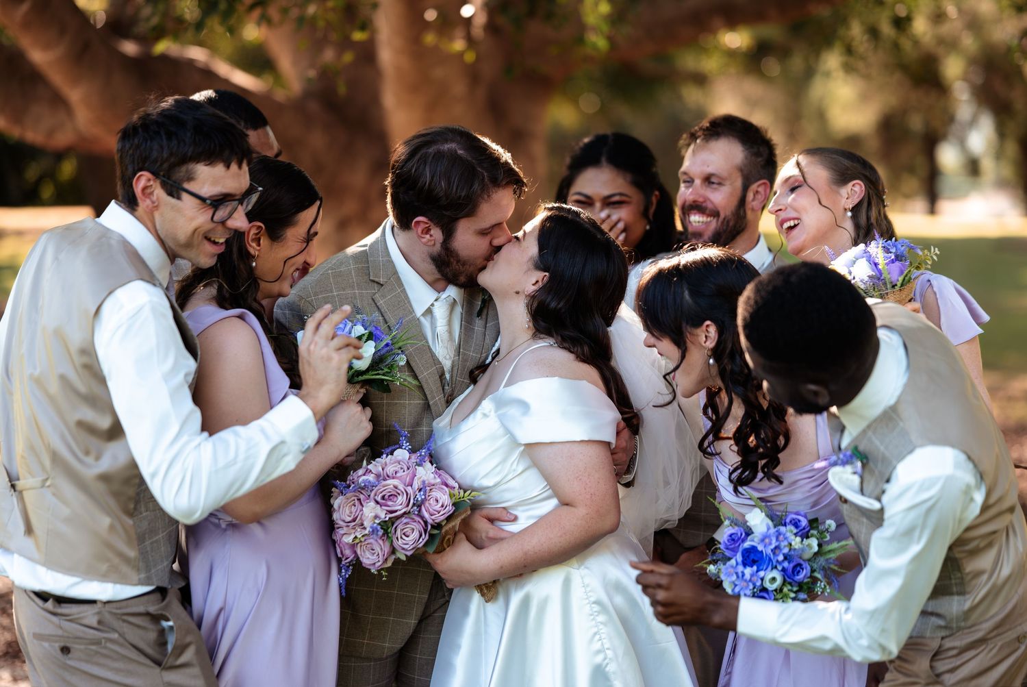 Wedding party celebrating as bride and groom kiss, surrounded by bridesmaids in lavender dresses holding bouquets outdoors.