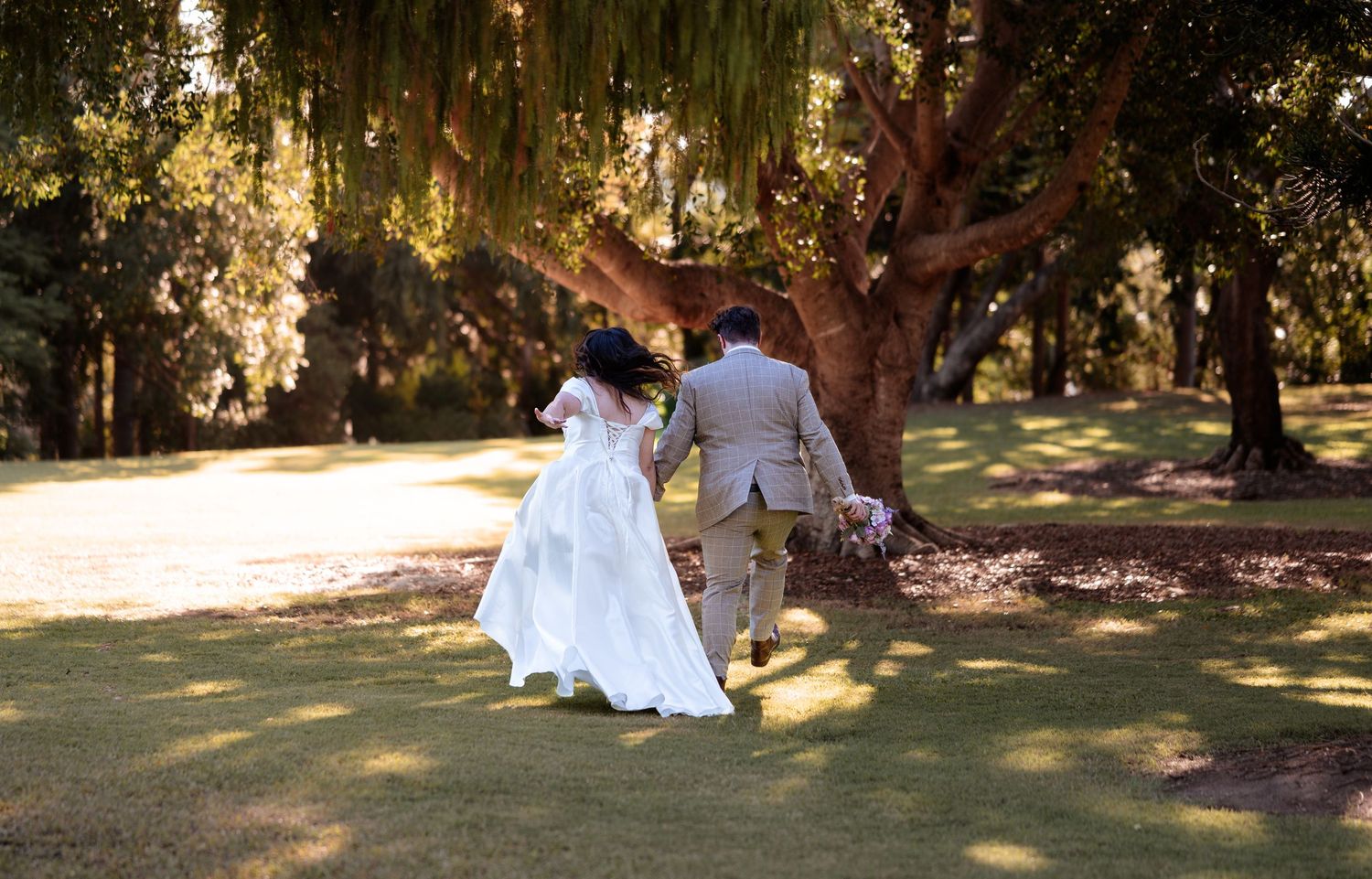 A bride and groom walk hand-in-hand through a lush green park beneath a large tree on their wedding day.