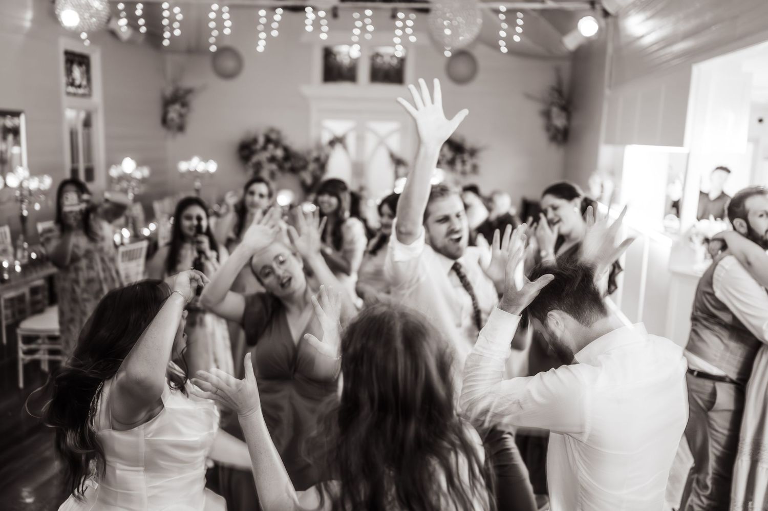 Black and white photo of wedding guests dancing joyfully with hands raised at a lively indoor reception.