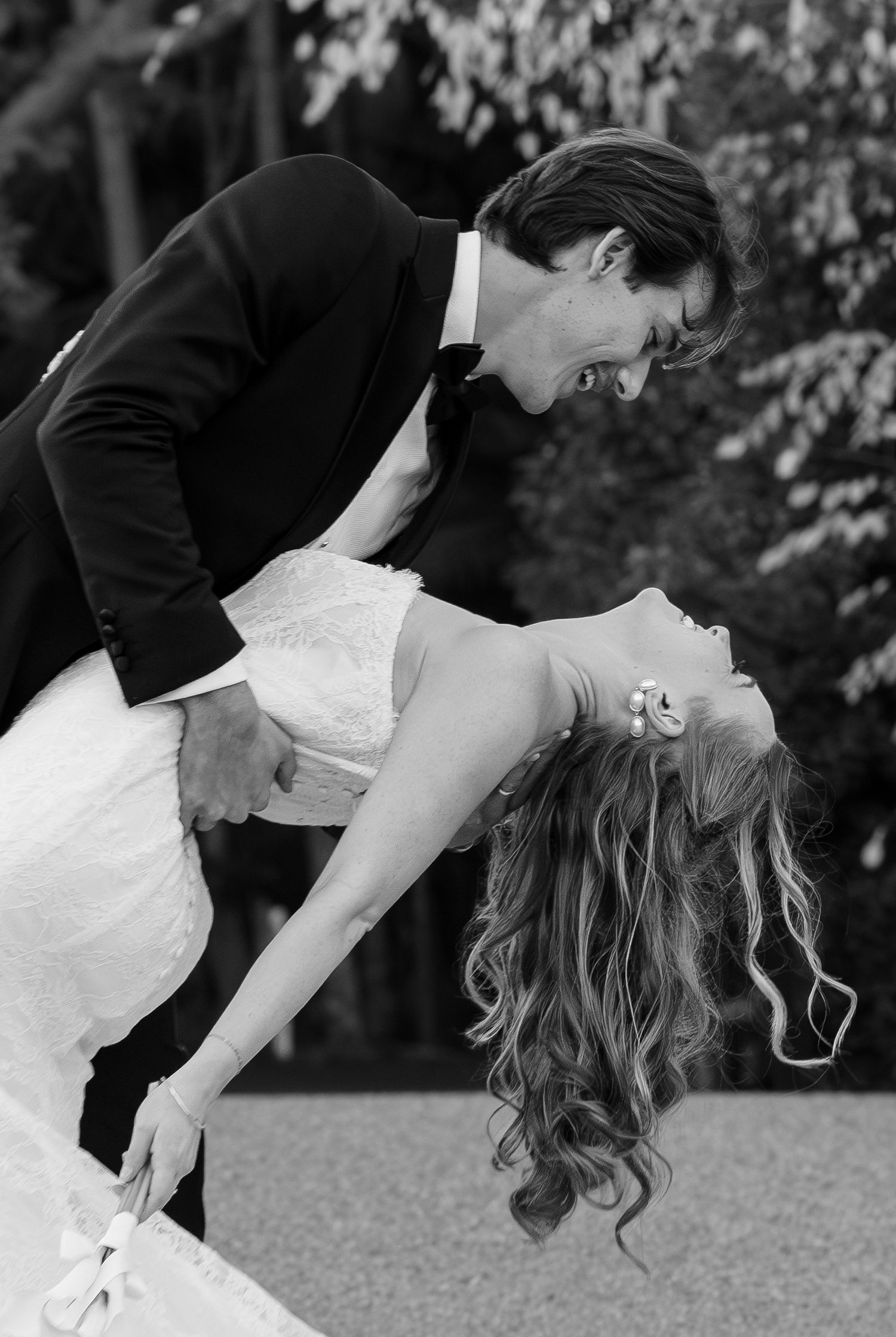 Black and white photo of groom dipping bride in elegant wedding attire, sharing a romantic moment outdoors.