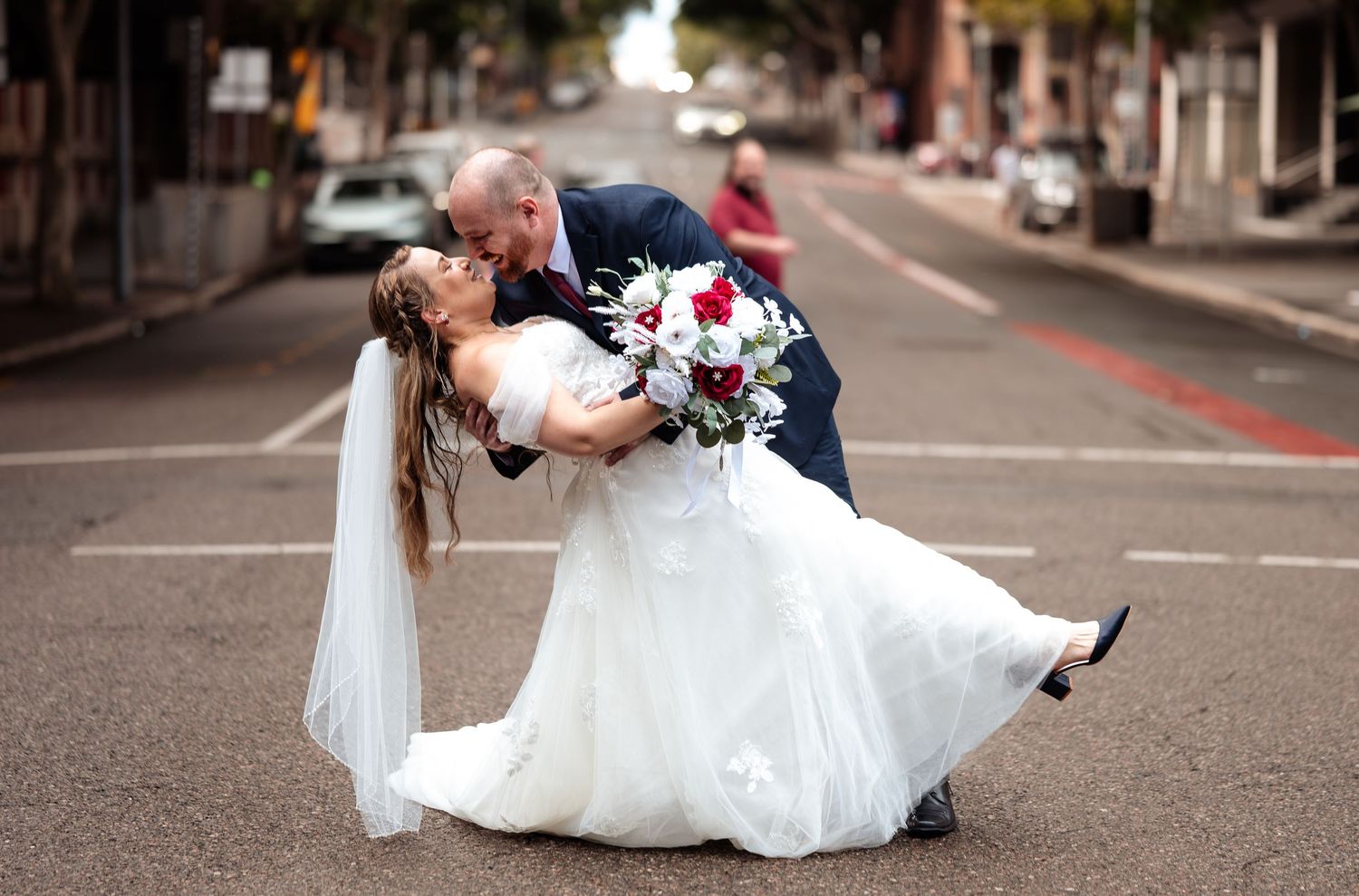 Bride and groom sharing a romantic dip kiss on a city street, bride holding a red and white bouquet.