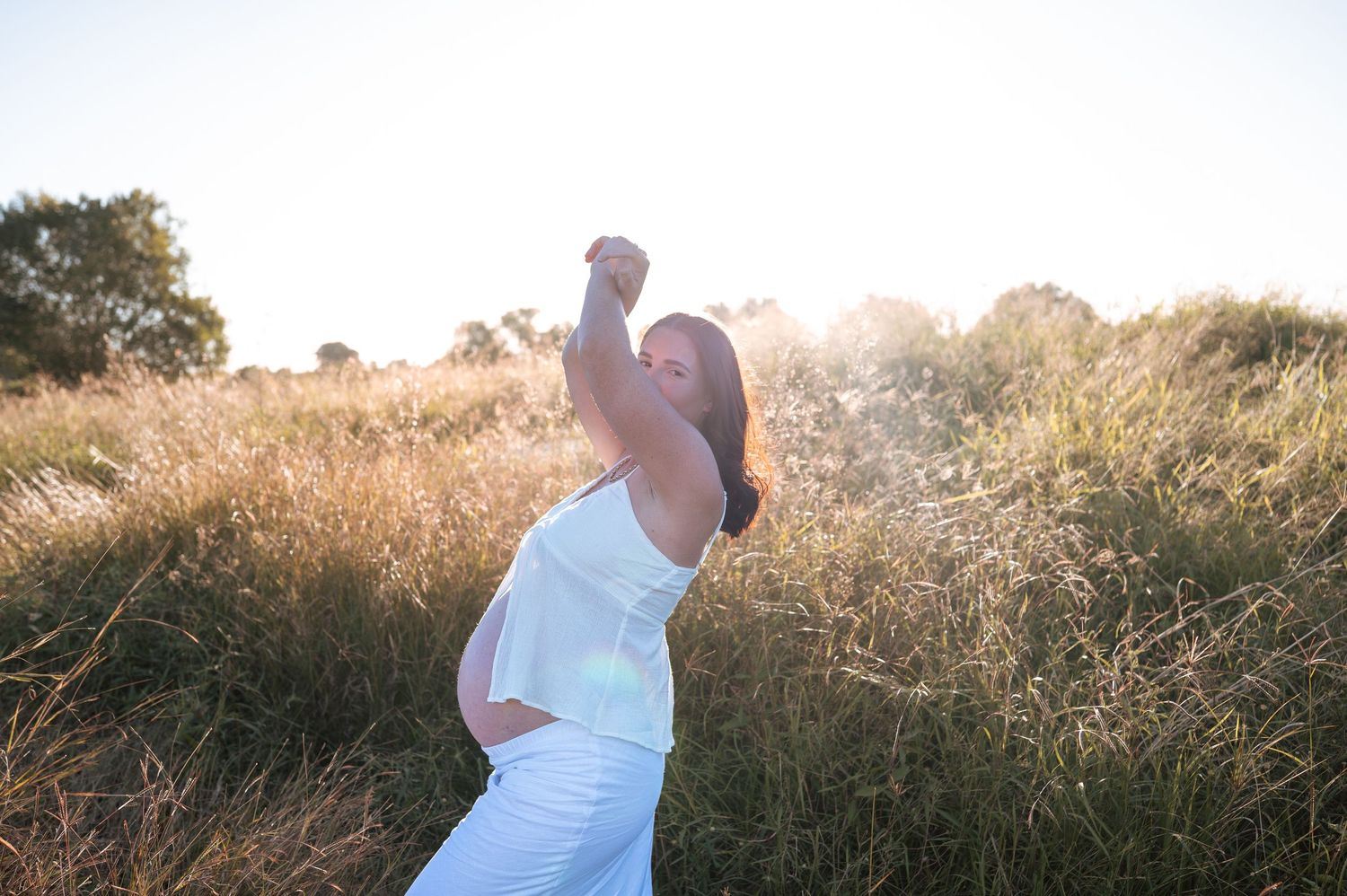 Maternity photoshoot in a field at sunset with woman in white dress.
