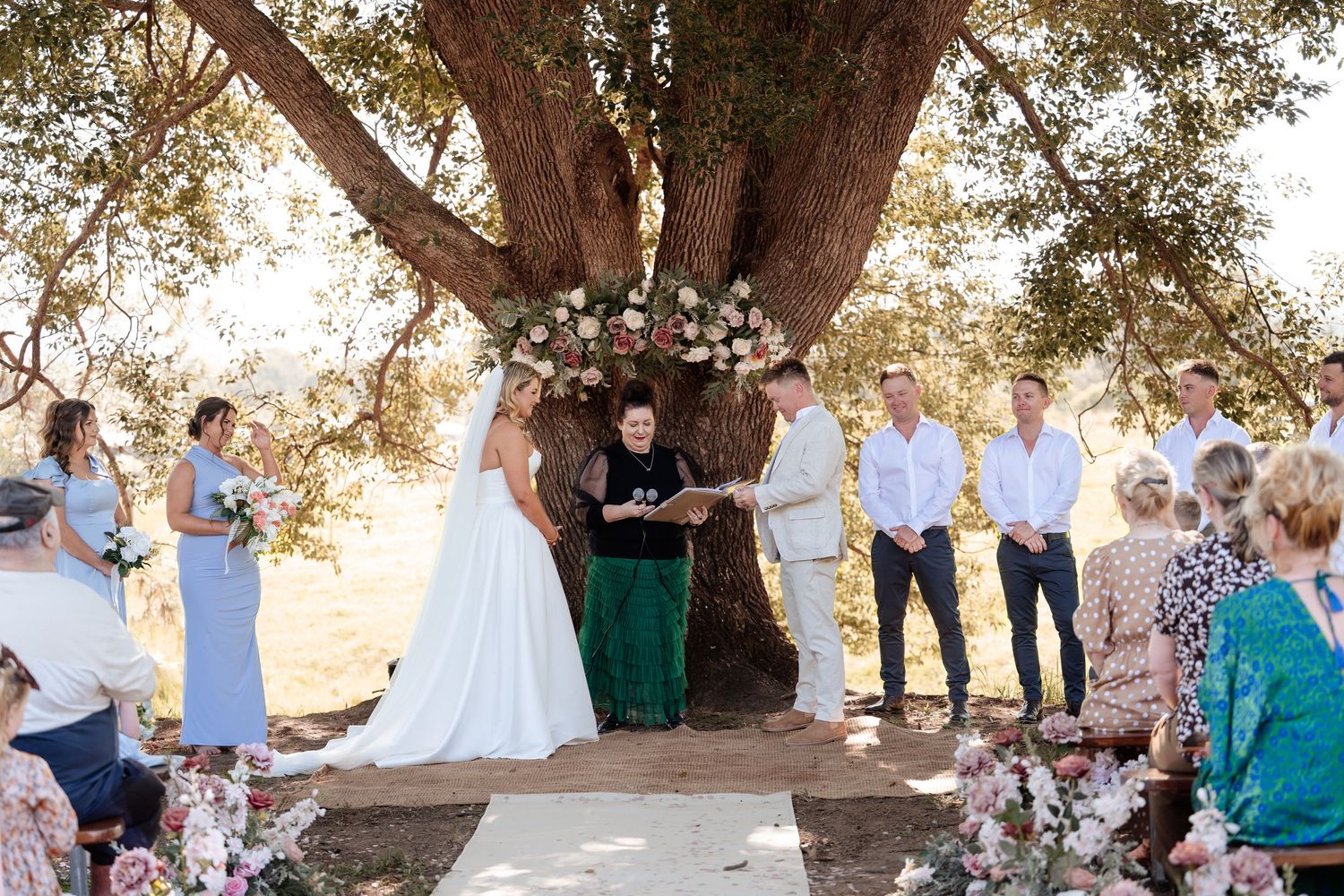 Bride and groom exchange vows under floral arch by ancient tree with guests watching.