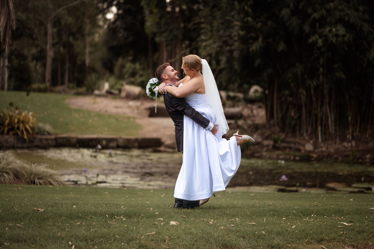 A bride in a flowing white dress holds a small child on a grassy lawn during a wedding photoshoot.
