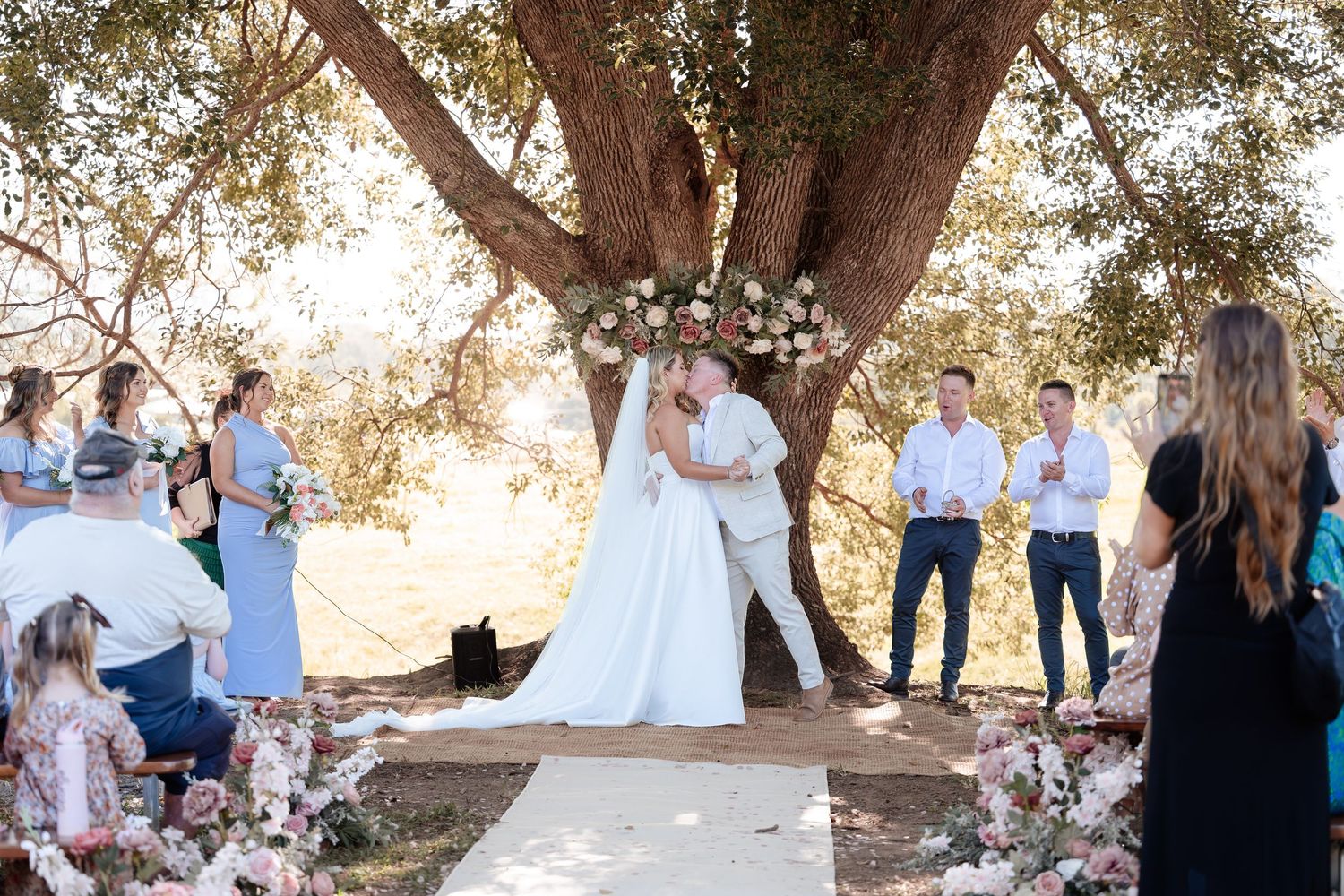 Couple sharing first kiss at outdoor wedding ceremony beneath a large tree with floral arch.