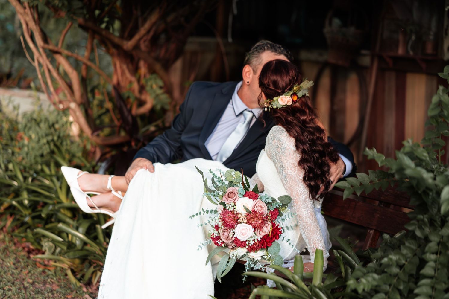 Bride and groom sharing a romantic kiss on a bench, bride holding a red and white floral bouquet.