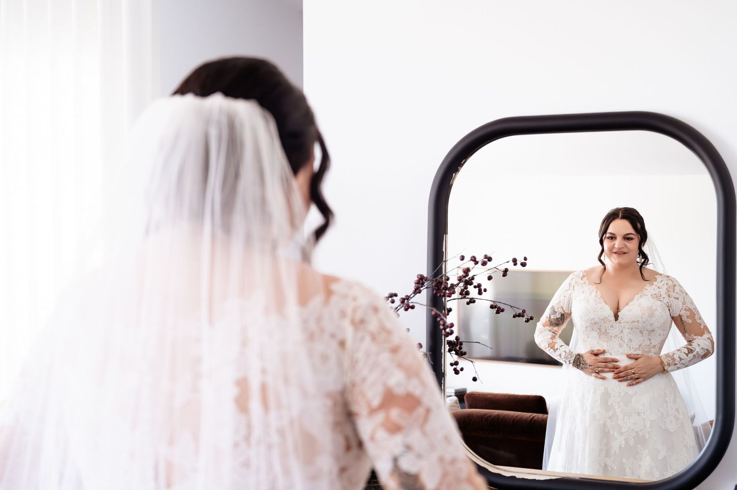 Bride in lace wedding gown and veil admires her reflection in a round mirror during bridal preparation.