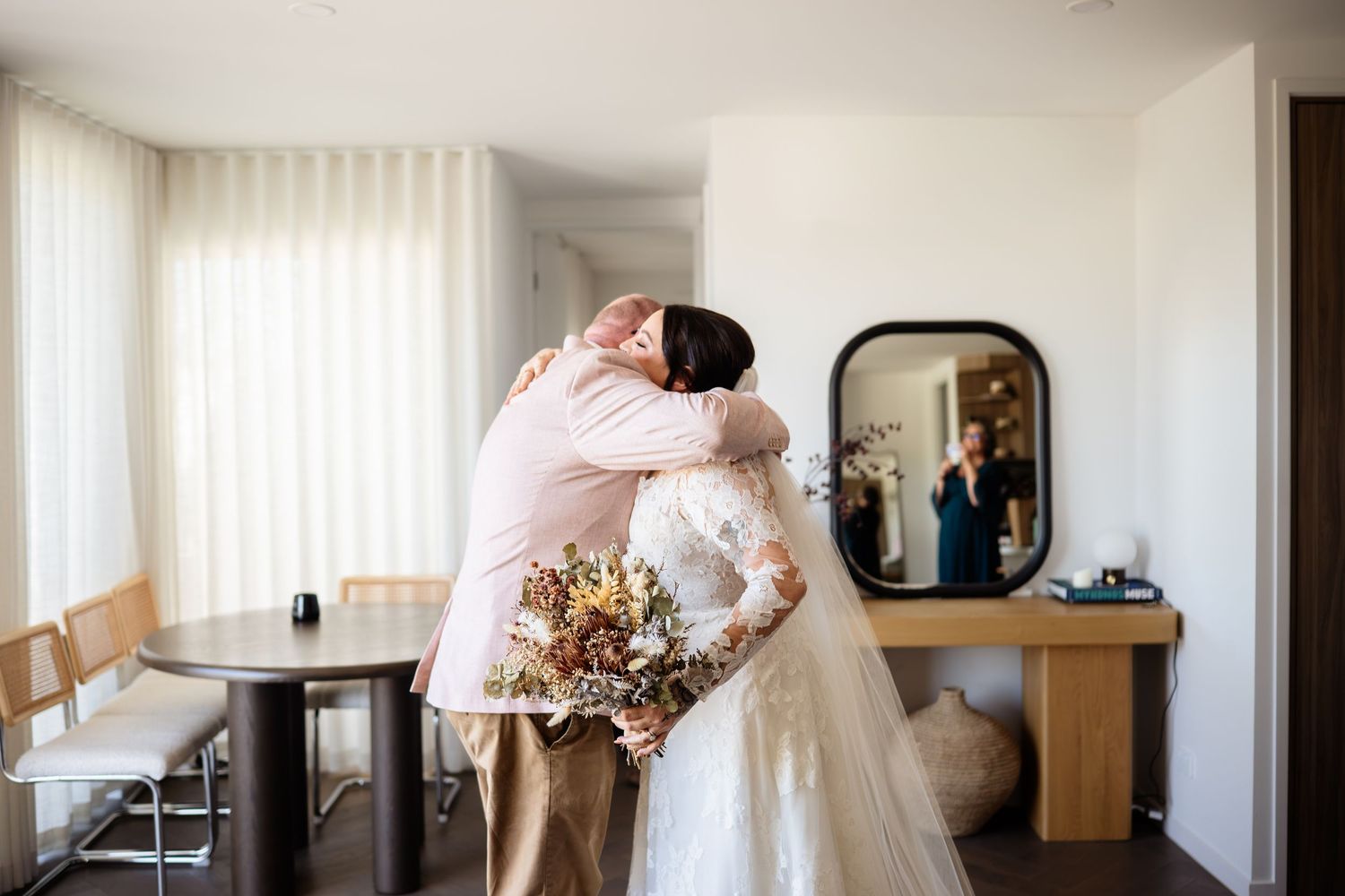 Bride in white lace gown hugging a guest while holding a dried floral bouquet in a bright hotel room.