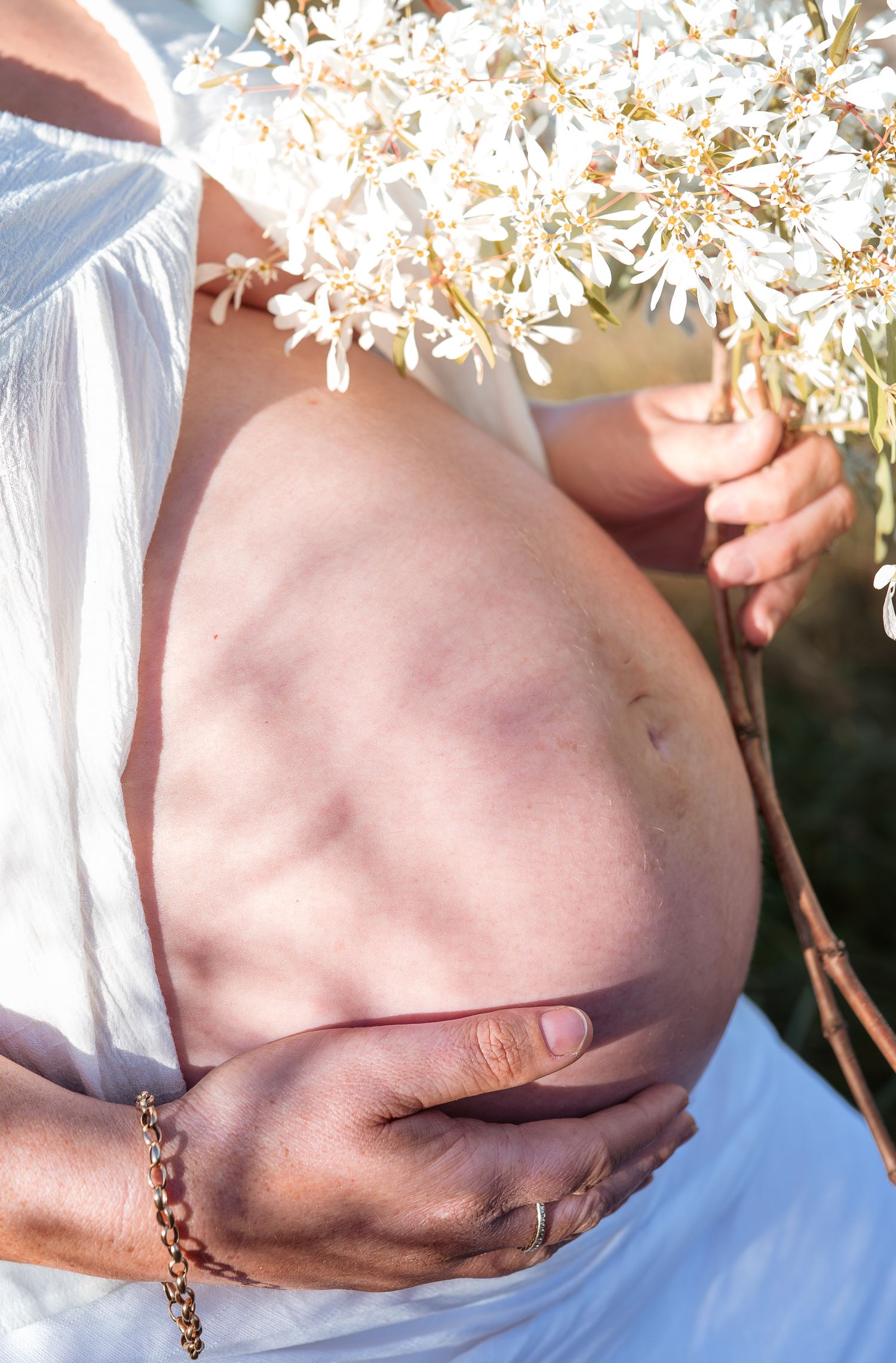 Close-up maternity photo showing a pregnant belly with white lace against a backdrop of delicate white flowers.
