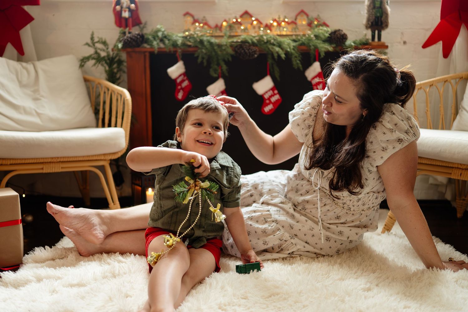 Mother and baby boy sitting on white rug enjoying Christmas decorations with stockings and garland fireplace.