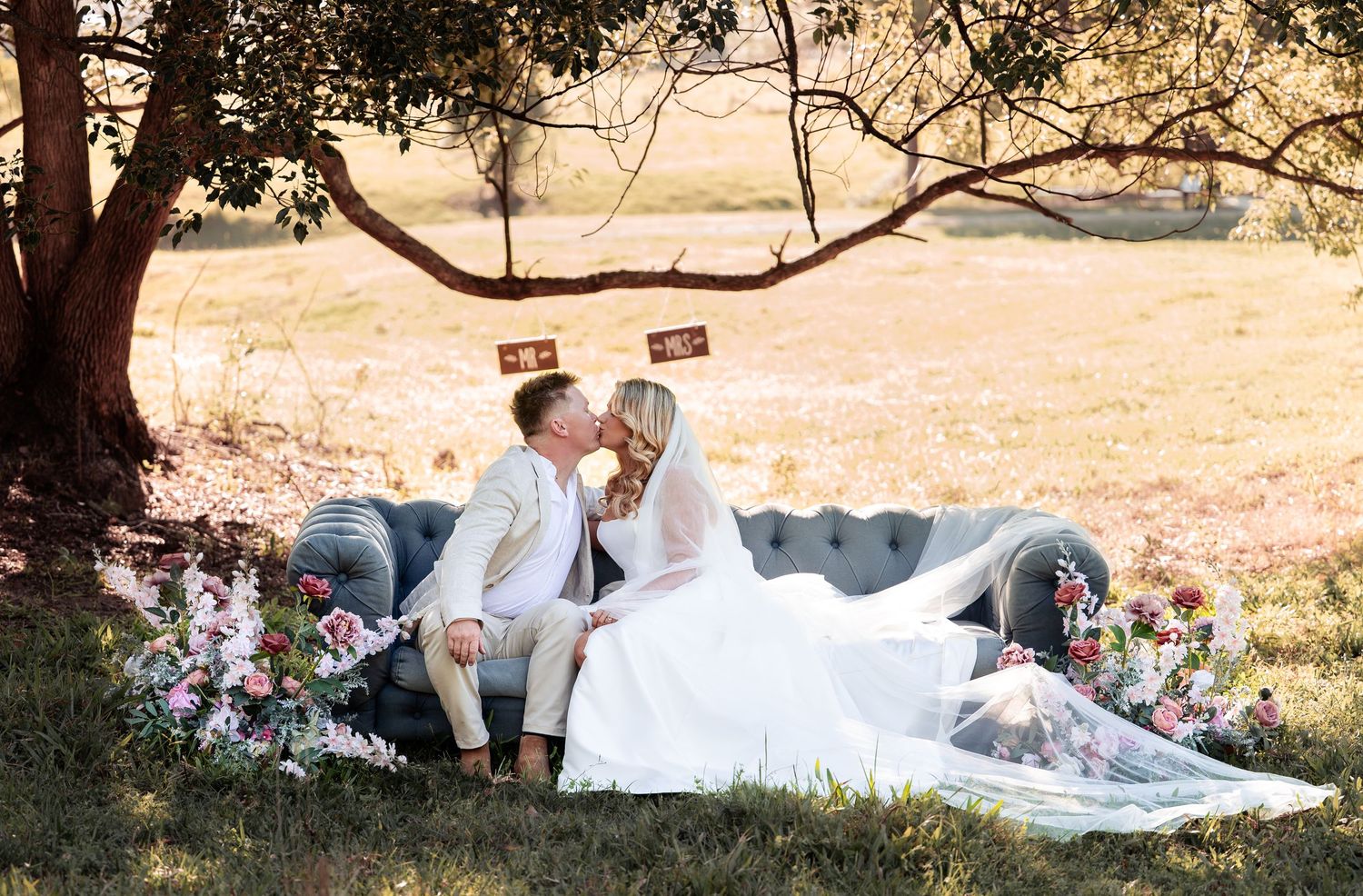 Bride and groom kissing on vintage sofa surrounded by flowers in outdoor setting.