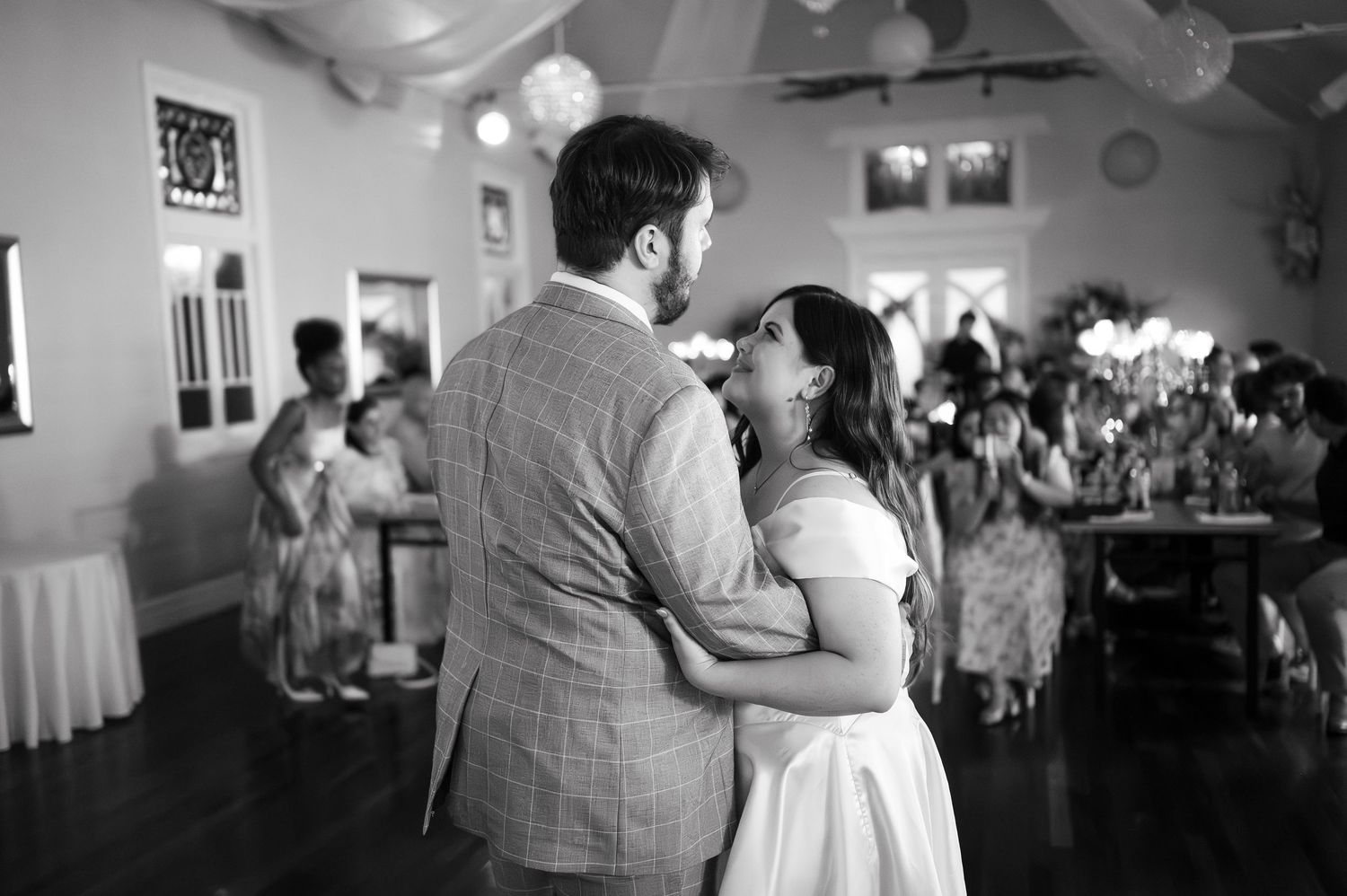 Black and white photo of a couple sharing their first dance at a wedding reception with guests watching in the background.