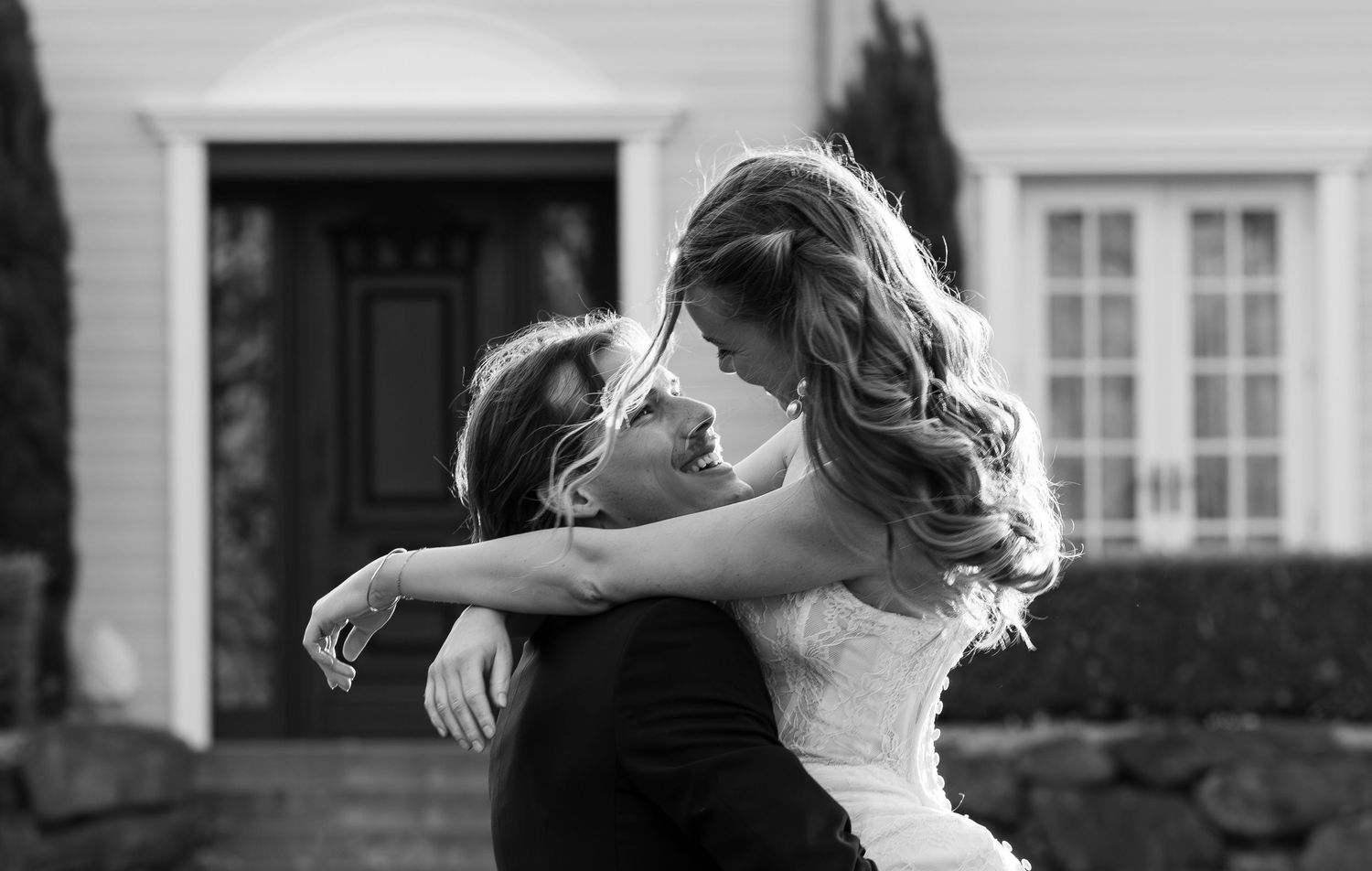 Bride and groom embrace in black and white wedding photo outside white house with windows and dark door.