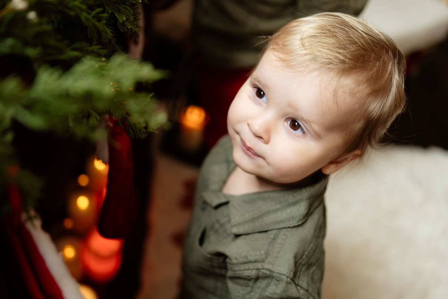 Cute toddler boy with blonde hair sitting near a decorated Christmas tree with festive lights and holiday decor.