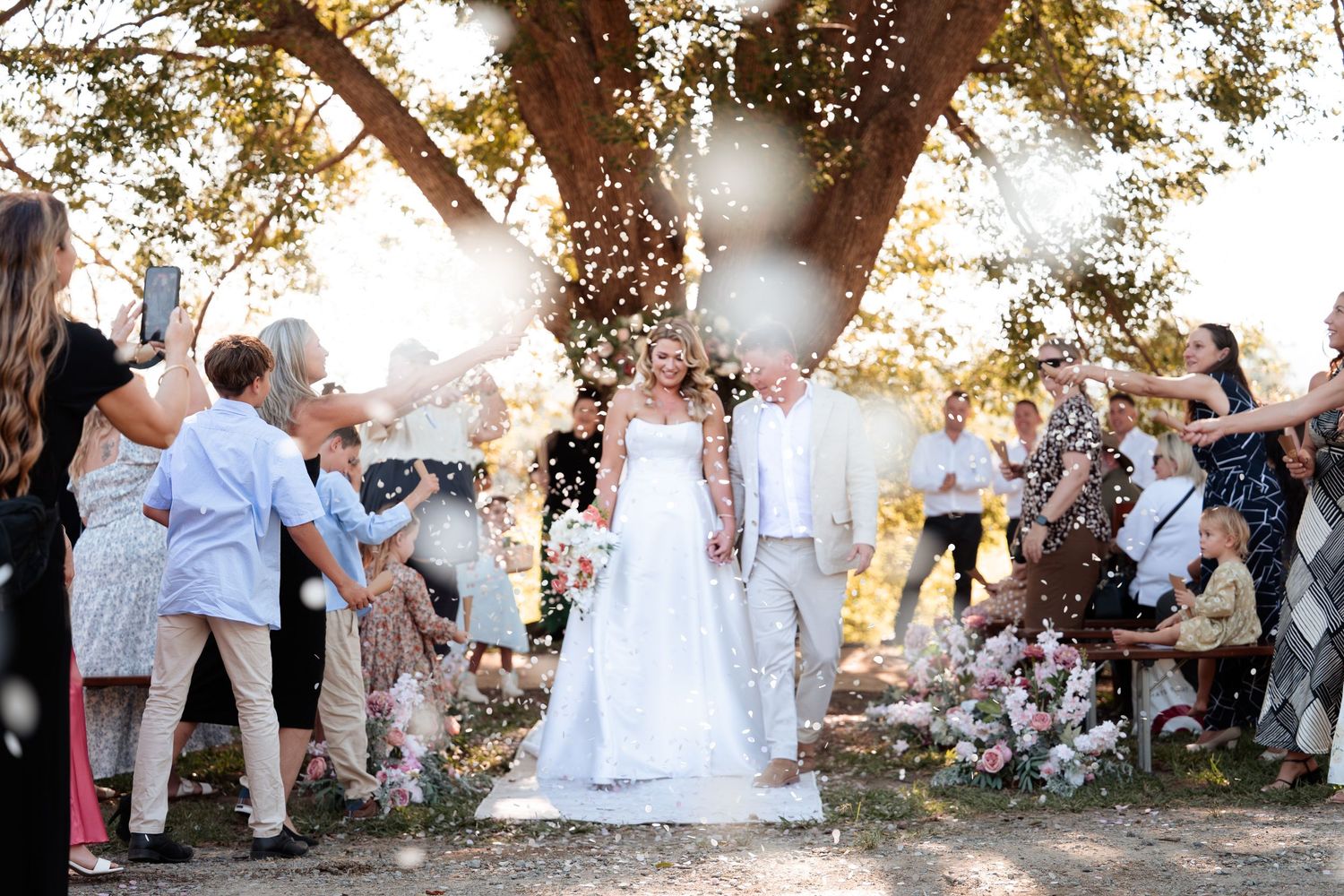 Bride and groom walk through confetti shower under oak tree surrounded by cheering wedding guests outdoors.