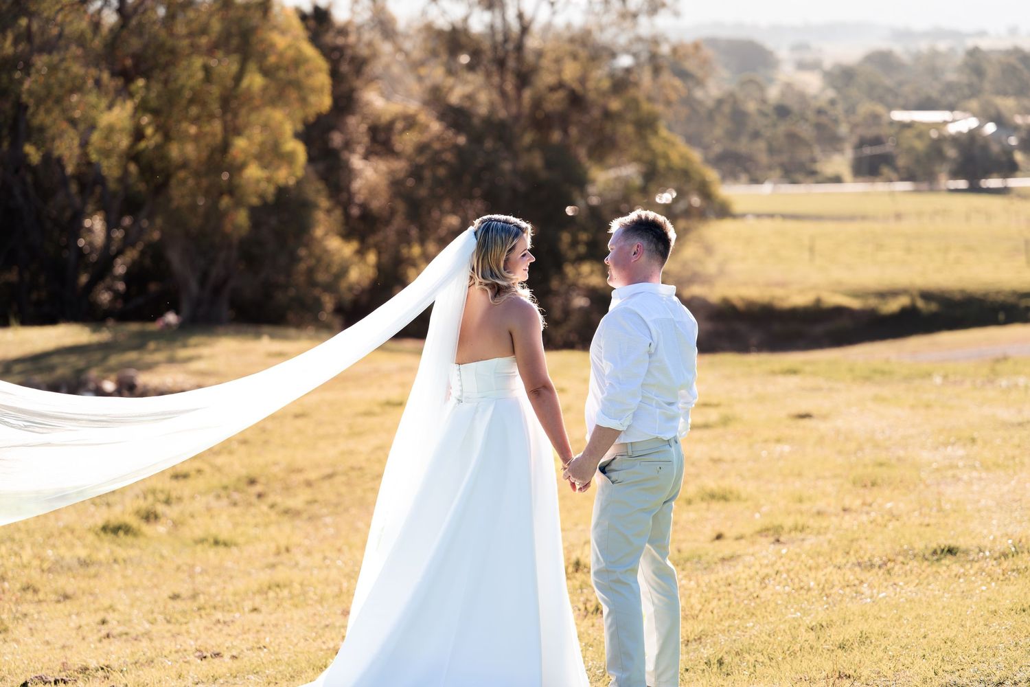 Bride and groom holding hands in a sunlit field, her veil flowing in the breeze, with rolling countryside in the background.
