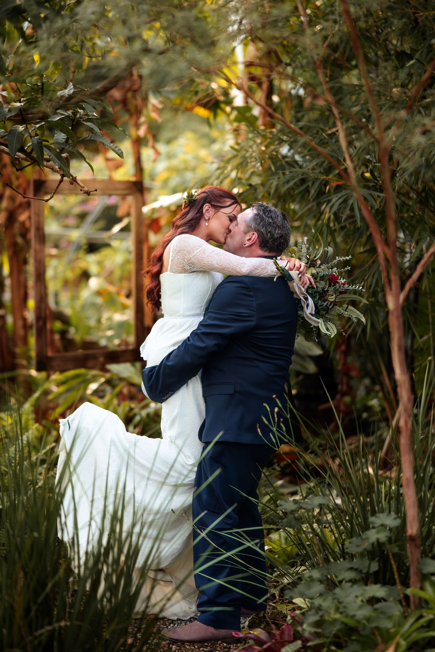 Groom lifts bride in white dress for a kiss surrounded by lush greenery in a romantic garden wedding photo.