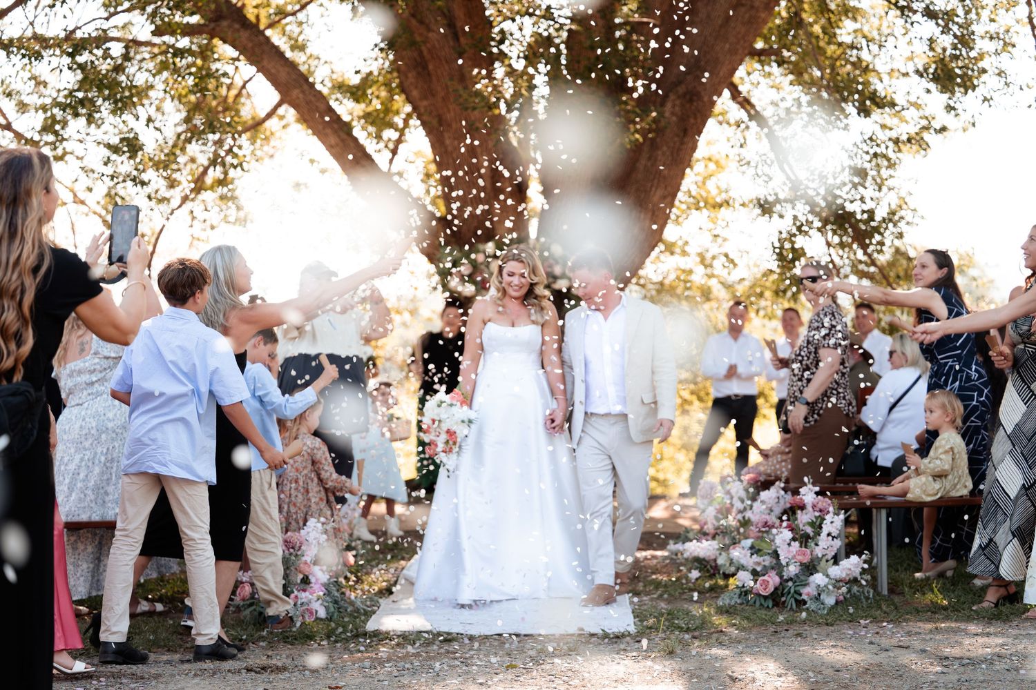 Bride and groom walk through confetti shower surrounded by cheering guests under a sunlit tree at outdoor wedding.