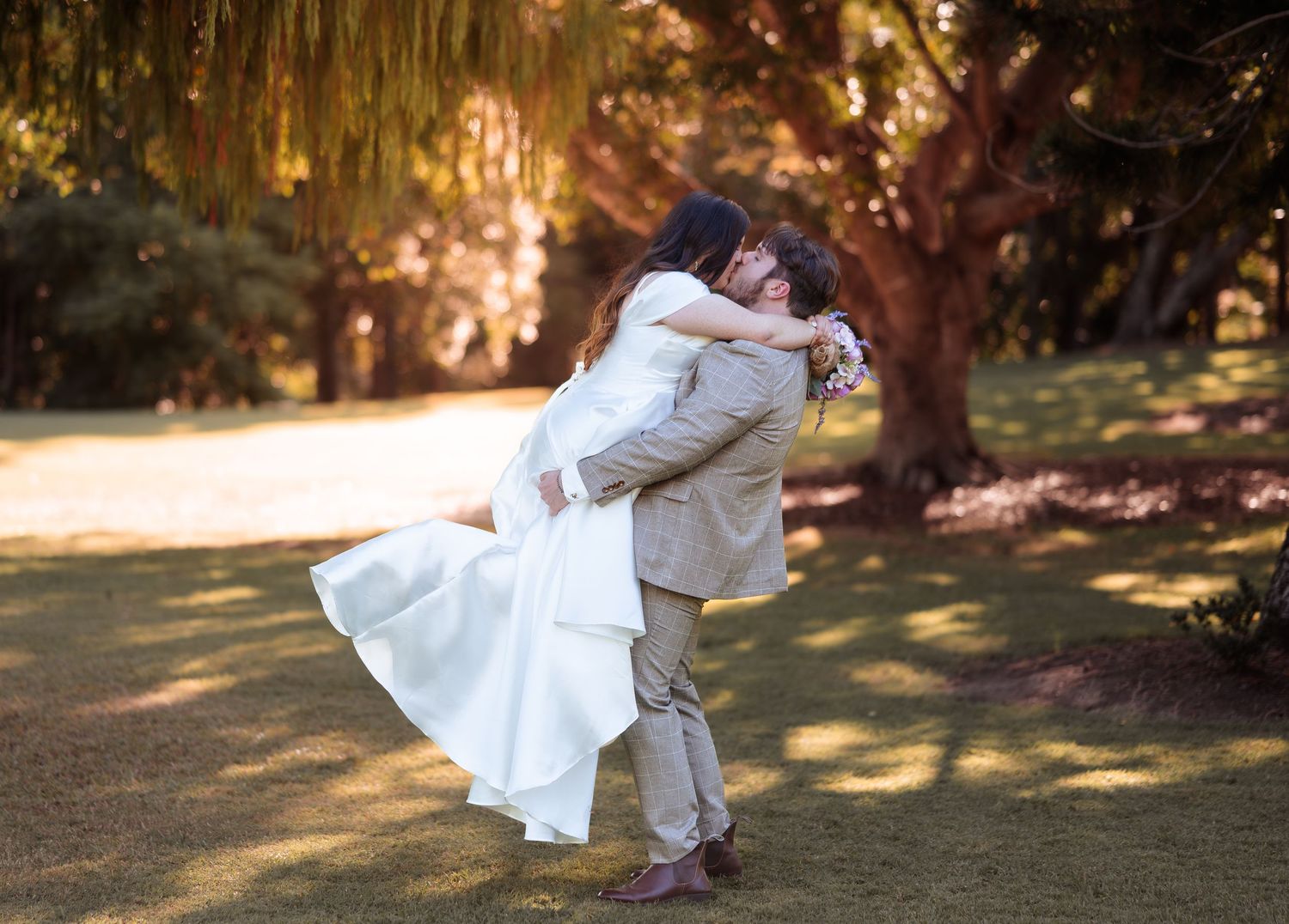 Groom lifting bride in a romantic embrace outdoors under lush green trees on wedding day.