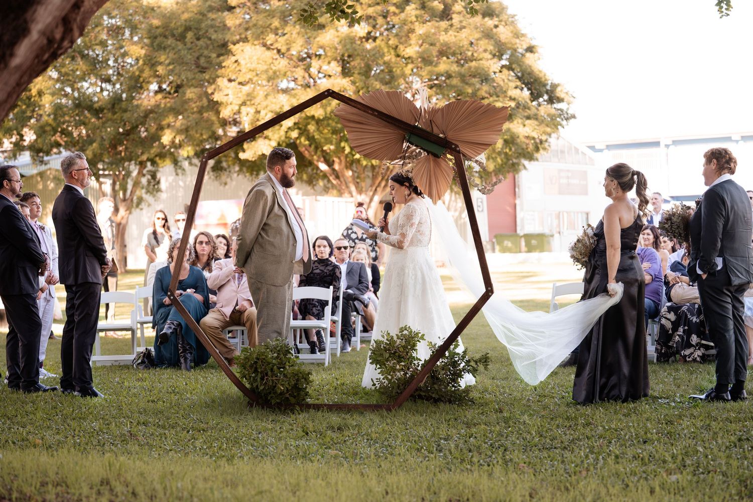 Outdoor wedding ceremony under geometric wooden arch on green lawn with guests seated in white chairs.