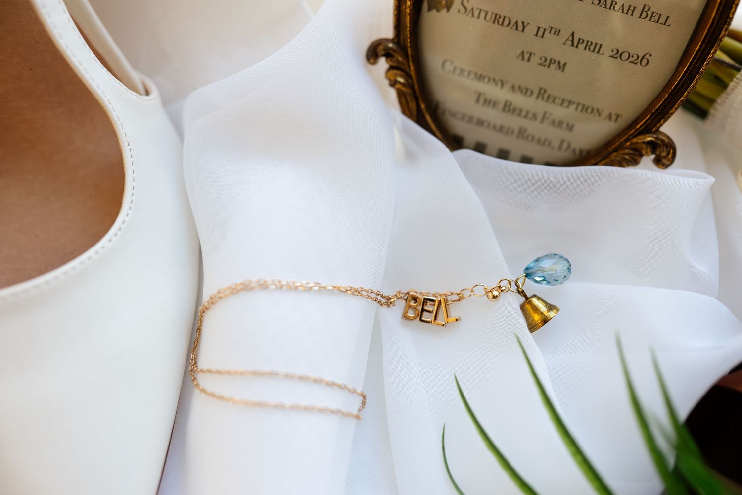 Gold name bracelet and charm bracelet displayed on white bridal fabric beside wedding invitation and mirror.
