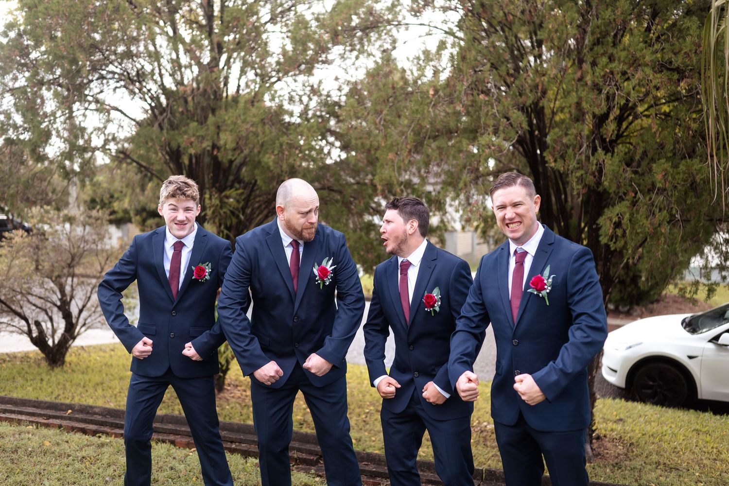 Four groomsmen in navy suits with red boutonnieres pose playfully flexing outside on wedding day.