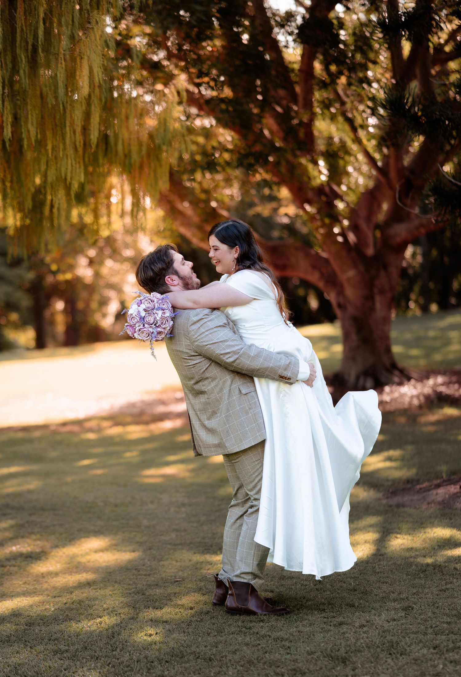 Groom lifts bride in white dress outdoors, surrounded by lush green trees and golden light, holding a purple bouquet.