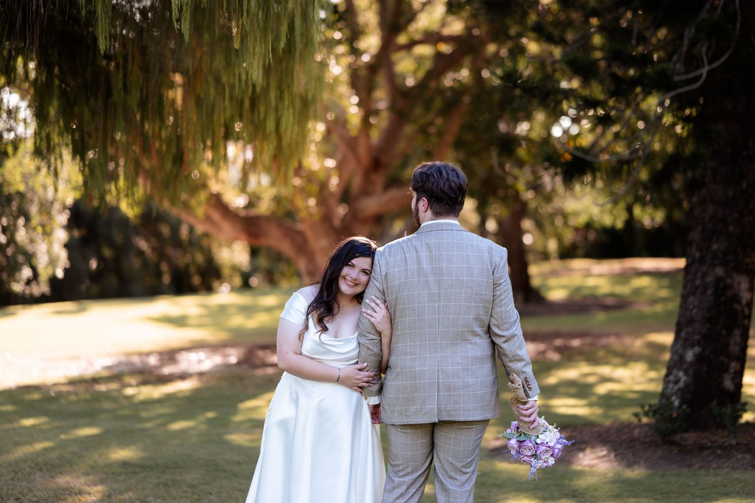 A bride and groom walk hand in hand through a lush garden, with the groom holding a purple bouquet.