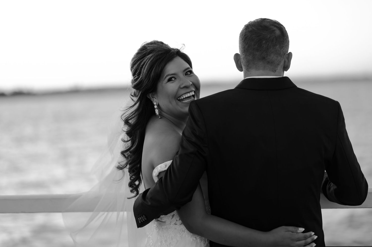 Black and white wedding photo of smiling bride in strapless gown embracing groom in suit near waterfront.
