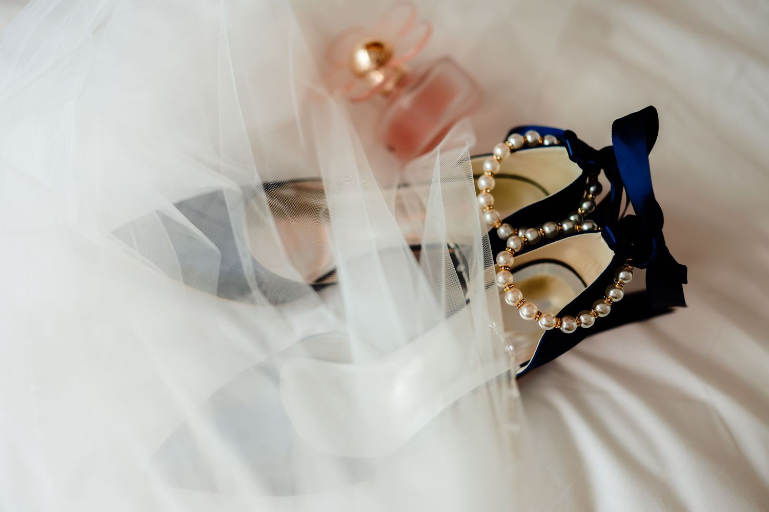 Elegant bridal accessories including pearl-embellished heels, blue satin shoes, and pink perfume bottle on white tulle veil.