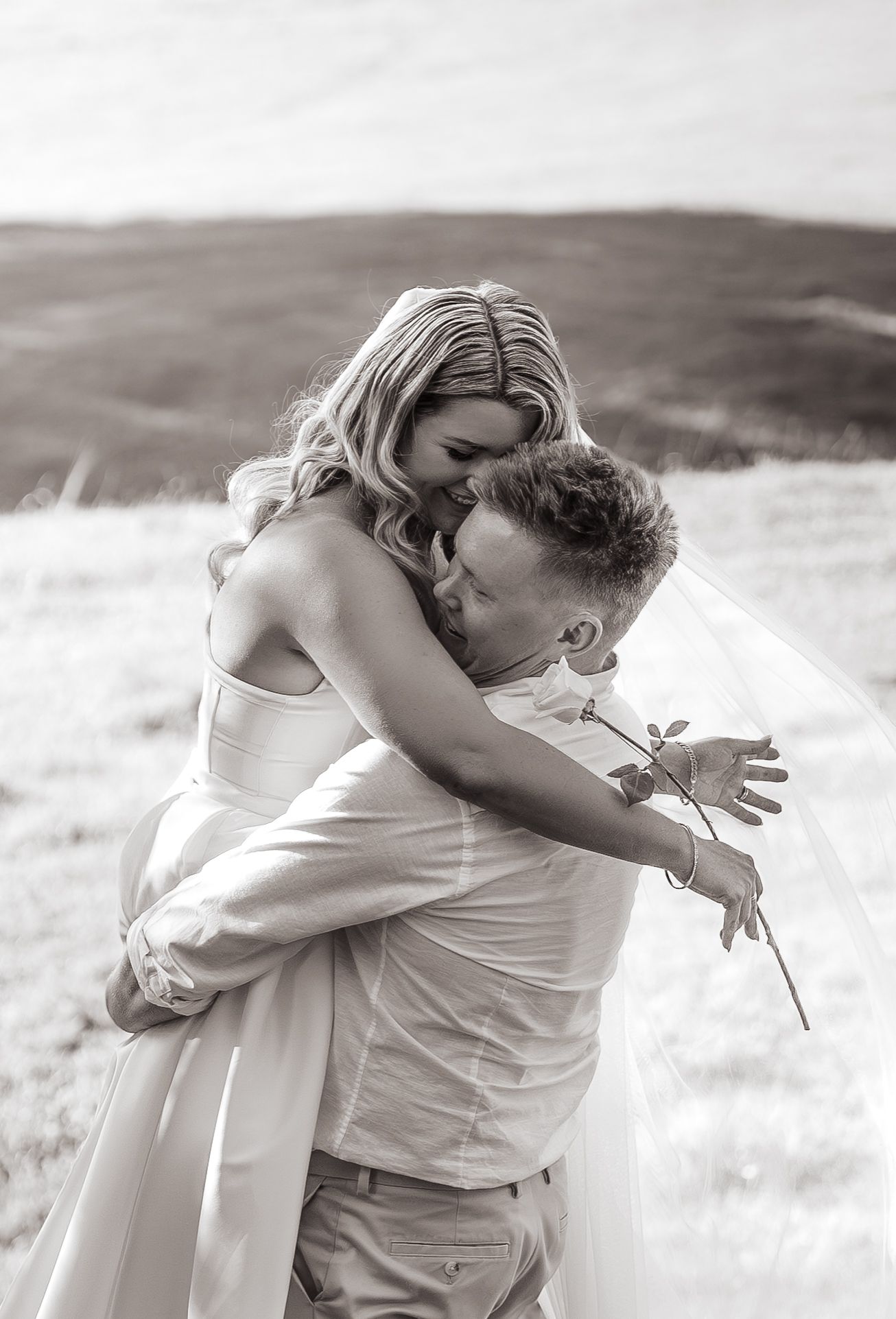 Black and white photo of a bride and groom embracing outdoors, she holds a single rose while wearing a wedding dress and veil.