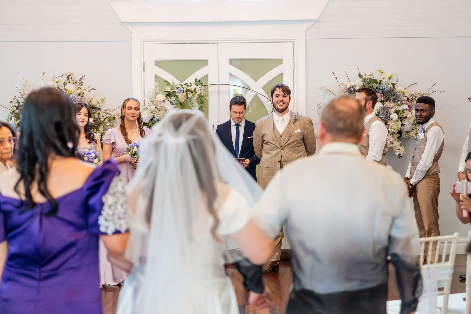 Bride walking down the aisle toward groom during indoor wedding ceremony with floral arrangements.