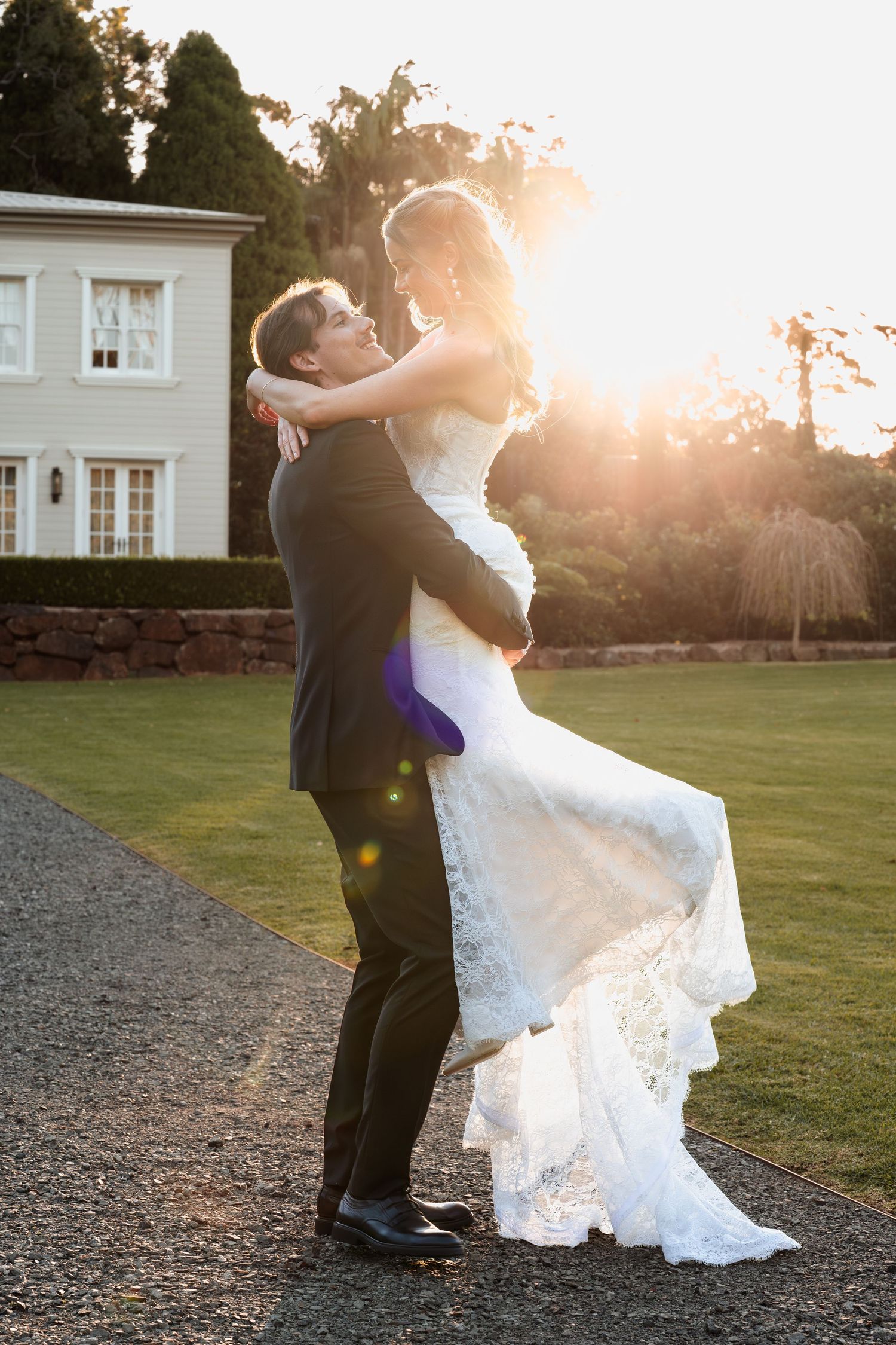 Bride and groom embrace on gravel driveway at sunset with elegant white house and manicured lawn in background.