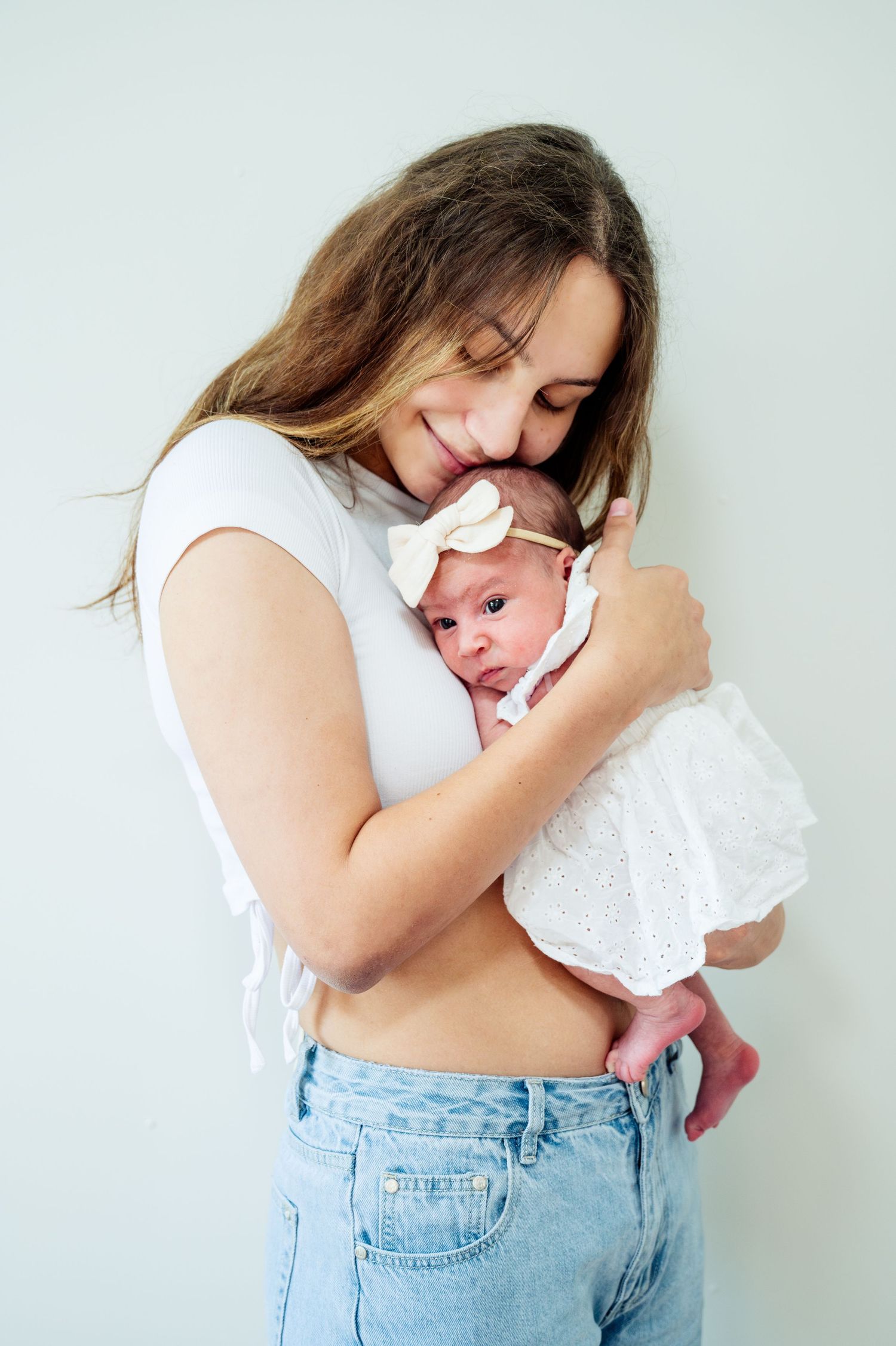 Mother in white shirt and denim cradles infant in gentle embrace against light background.