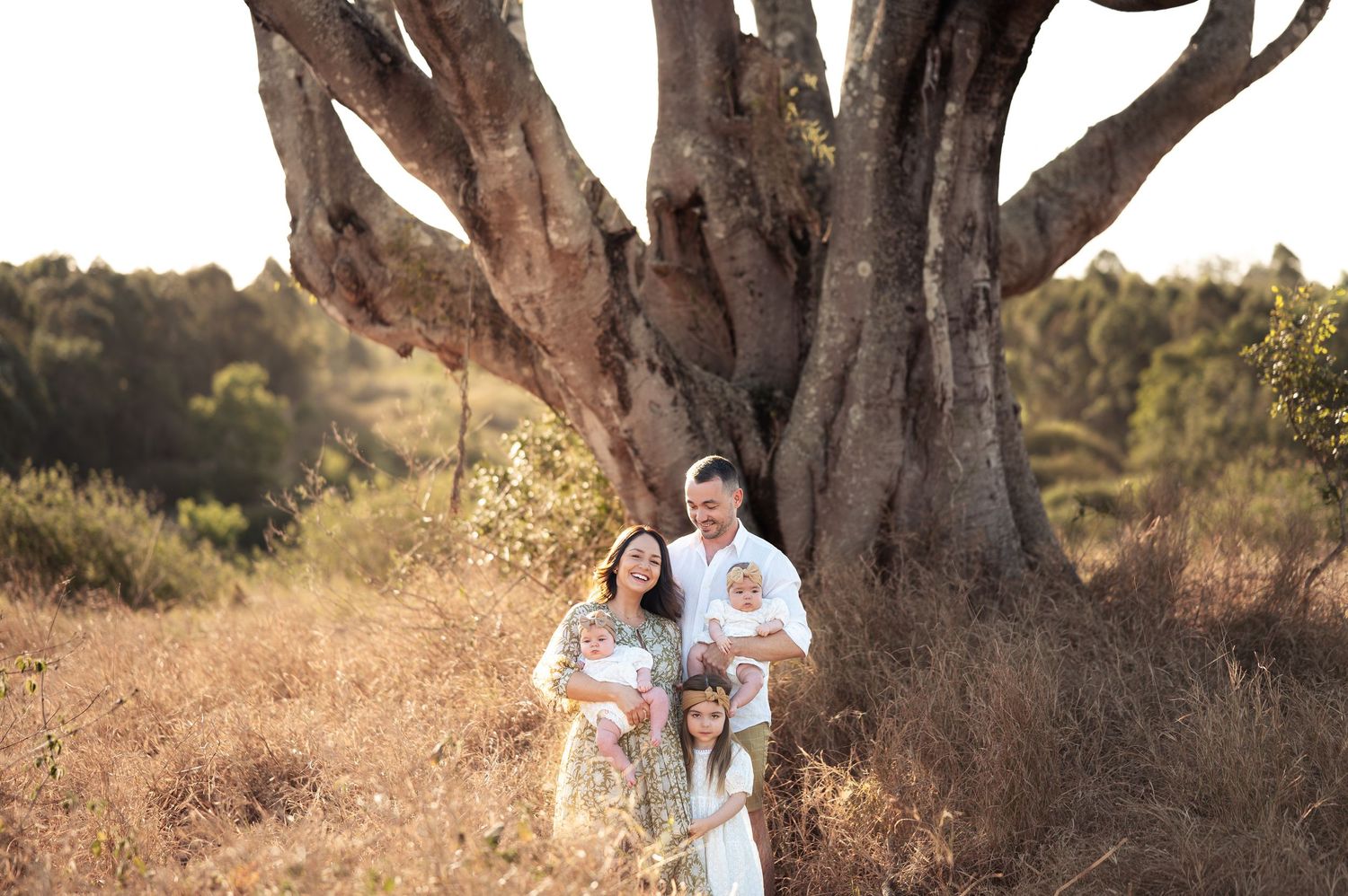 Family photoshoot under large trees with golden sunset lighting in background.