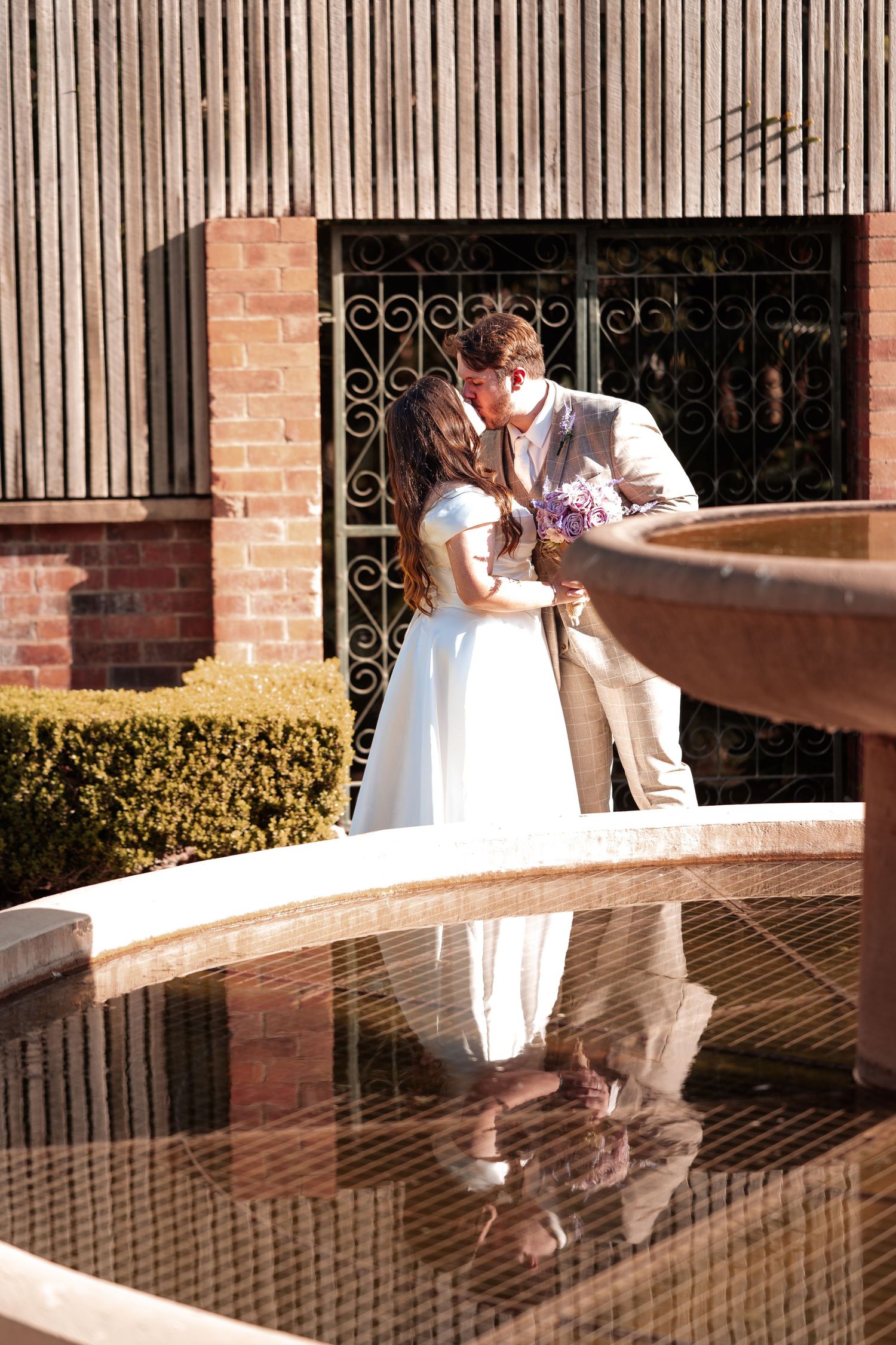 Bride and groom sharing a kiss beside a reflective fountain, with brick wall and ornate iron gate backdrop.
