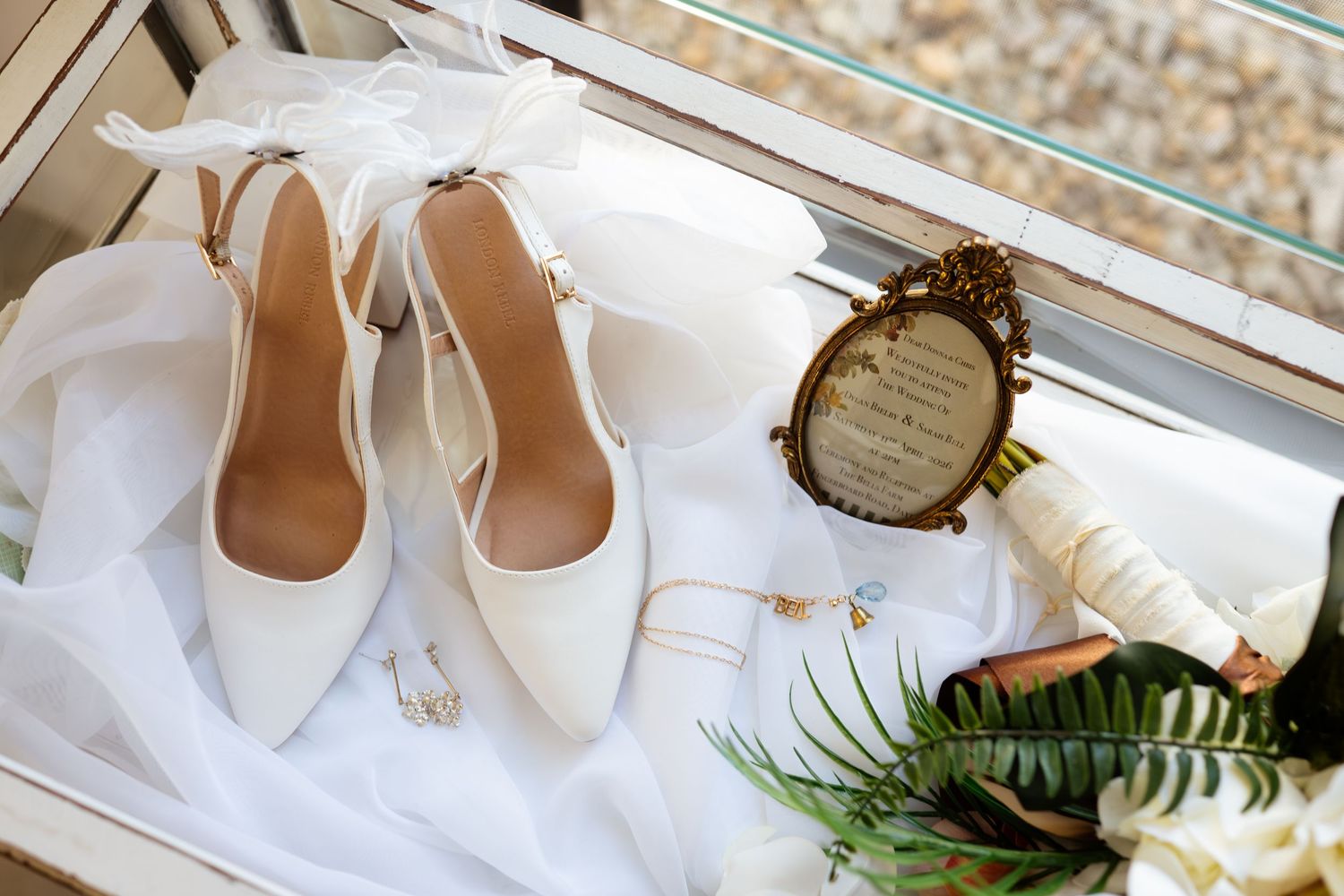 White bridal heels with ribbon ties displayed alongside gold jewelry, a vintage mirror, and a floral bouquet on white fabric.