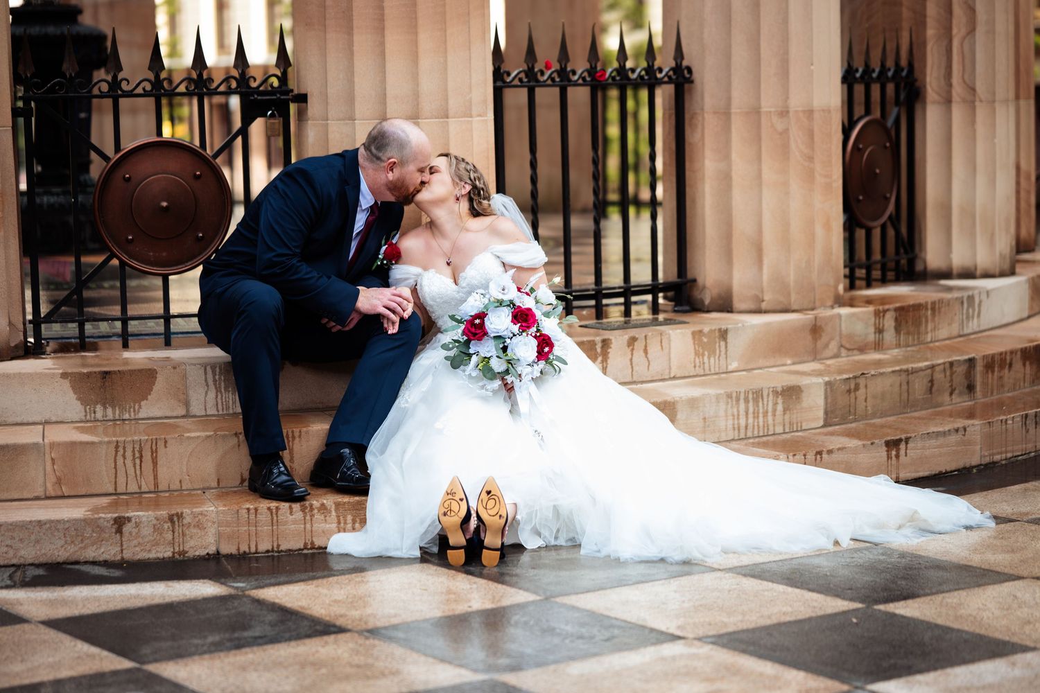 Bride and groom share a romantic kiss on stone steps, wedding dress spread out, gold heels placed nearby.