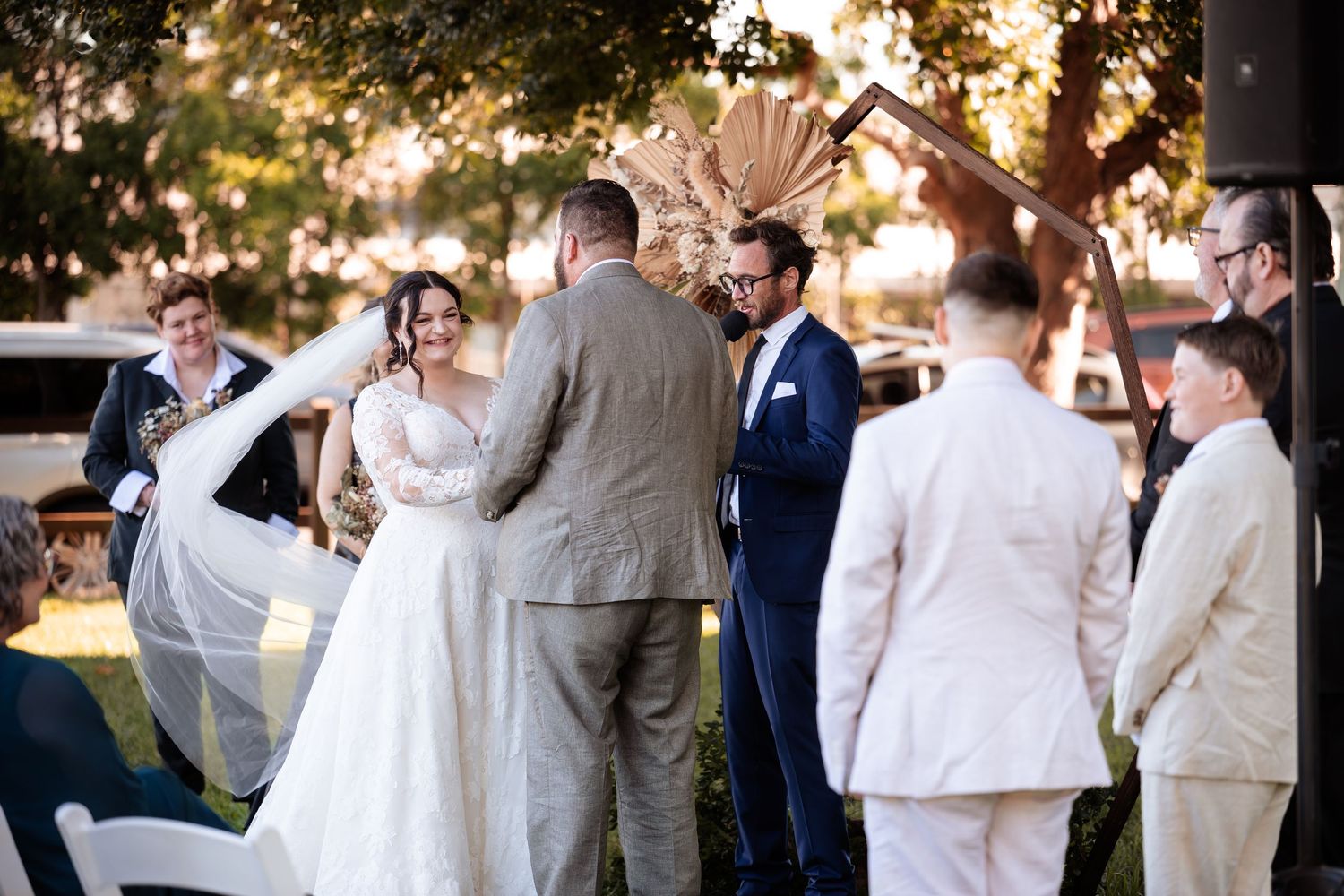 Outdoor wedding ceremony with bride in lace gown and groom in navy suit exchanging vows under decorative arch.