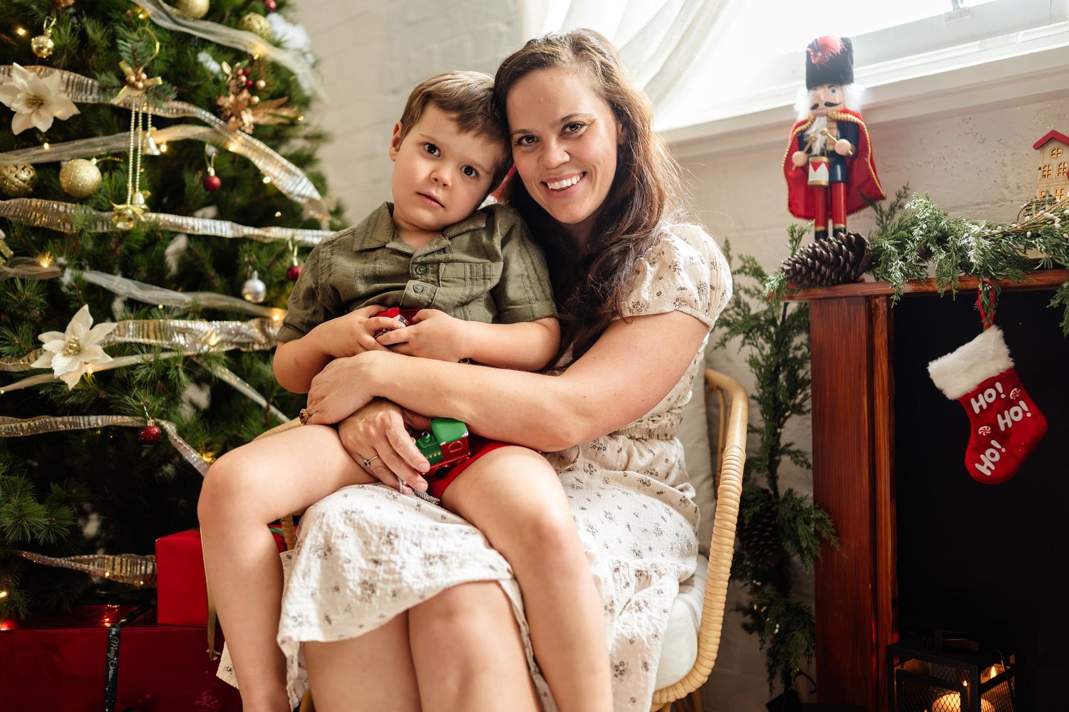 Woman and young boy smiling together by a decorated Christmas tree with holiday ornaments and gifts.