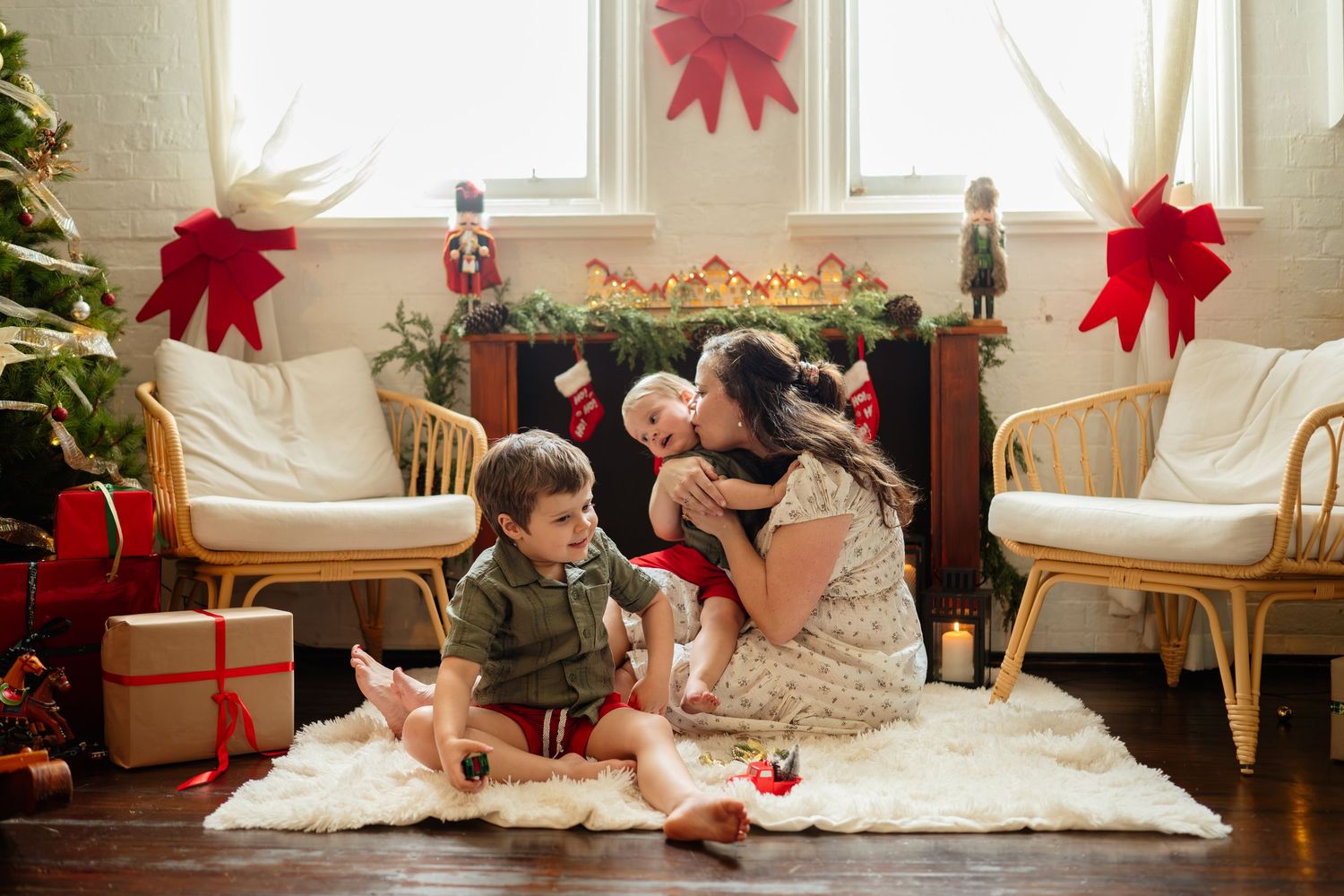 Mother and two children sitting on a white fur rug in a festively decorated Christmas living room with red bows.