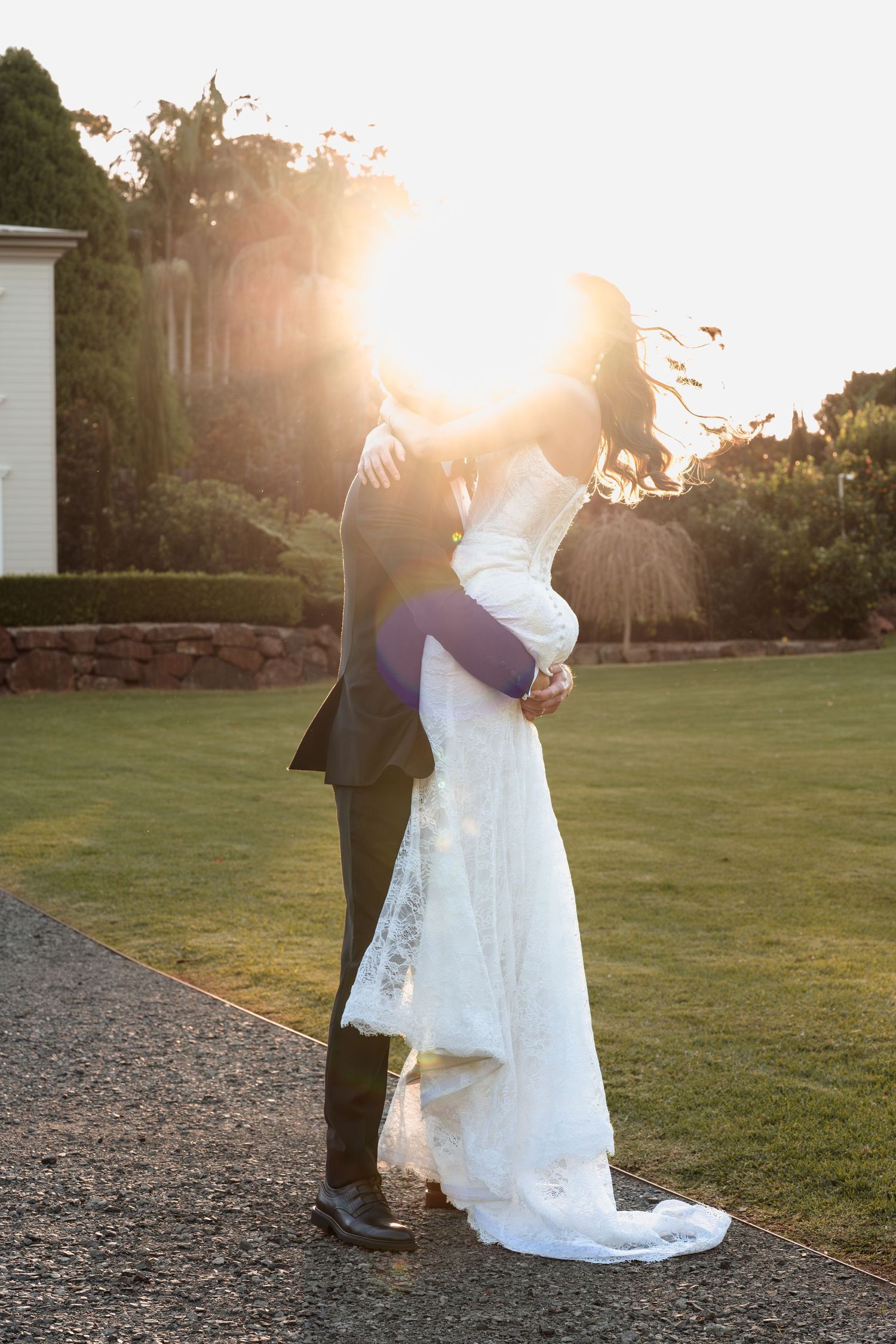Bride and groom embrace on garden pathway at sunset with dramatic sun flare creating romantic wedding portrait.