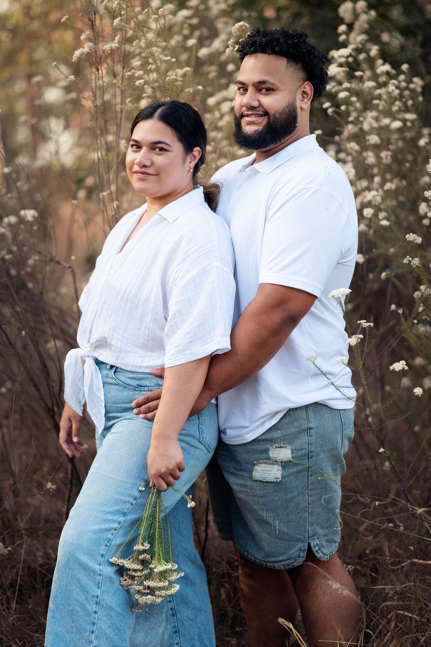 Couple in matching white shirts and denim posing together among wildflowers.